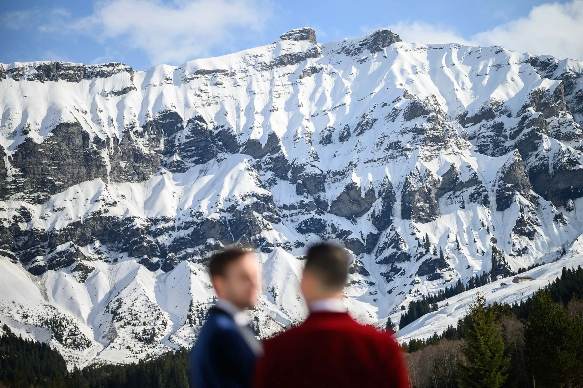 Deux hommes flous en costume discutent devant une montagne enneigée avec des pics rocheux, forêt de conifères en premier plan.