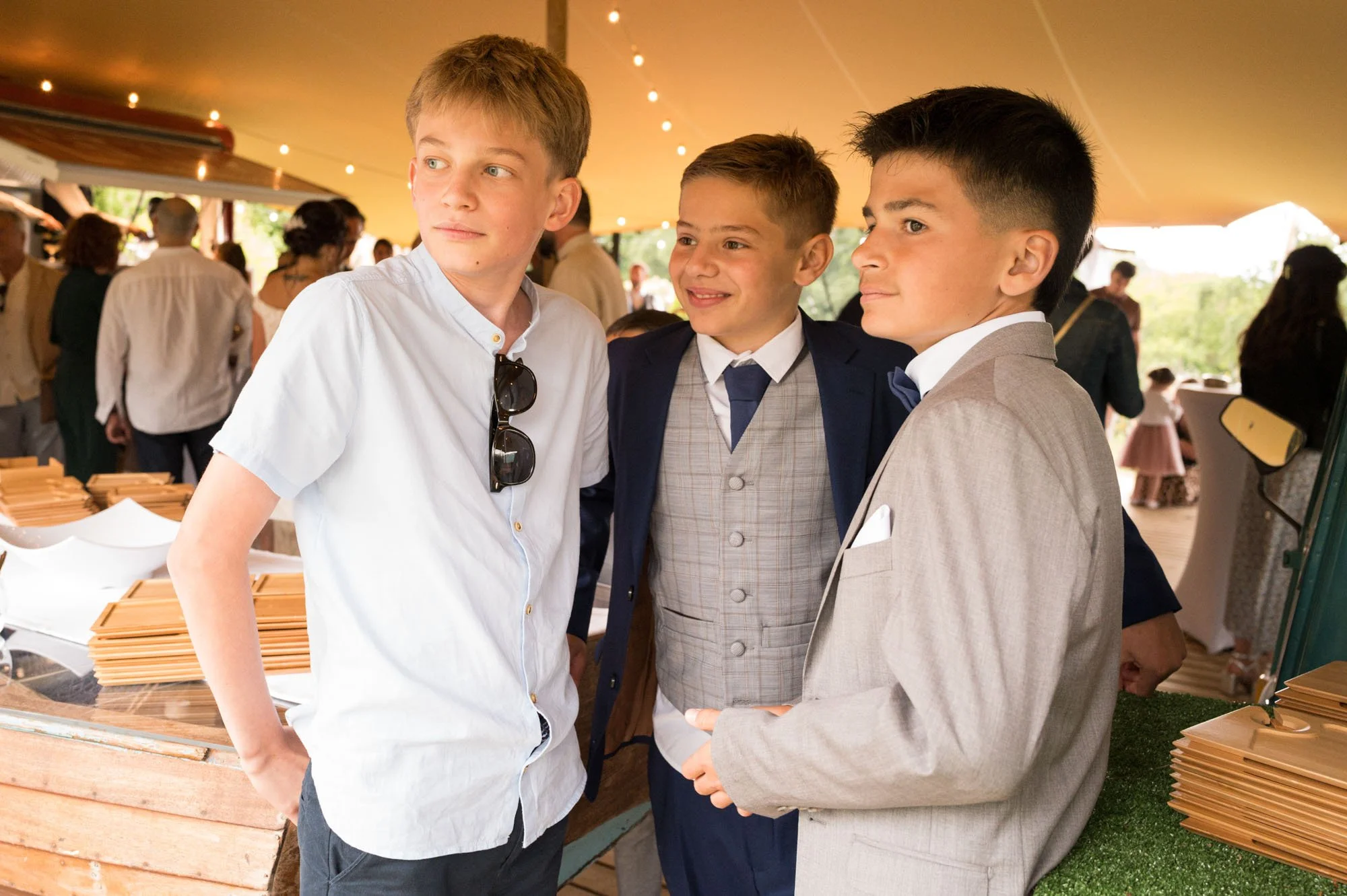 Trois jeunes garçons élégamment habillés à un évènement, se tenant sous une tente avec des invités en arrière-plan.
