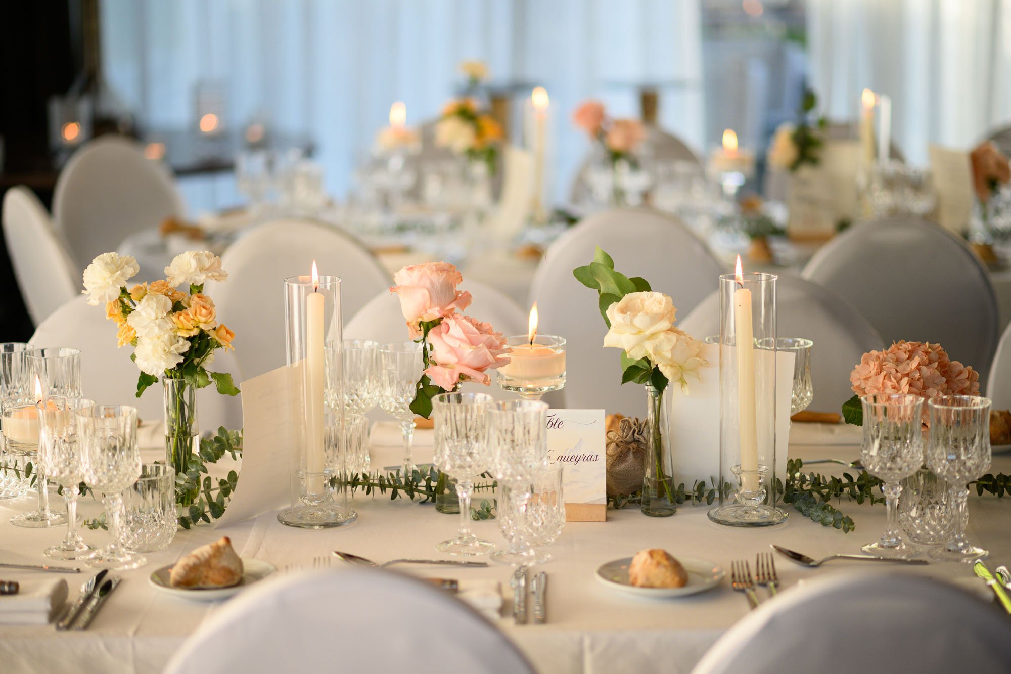Table de mariage élégamment décorée avec des fleurs pastel, bougies et verre à vin, en intérieur lumineux.