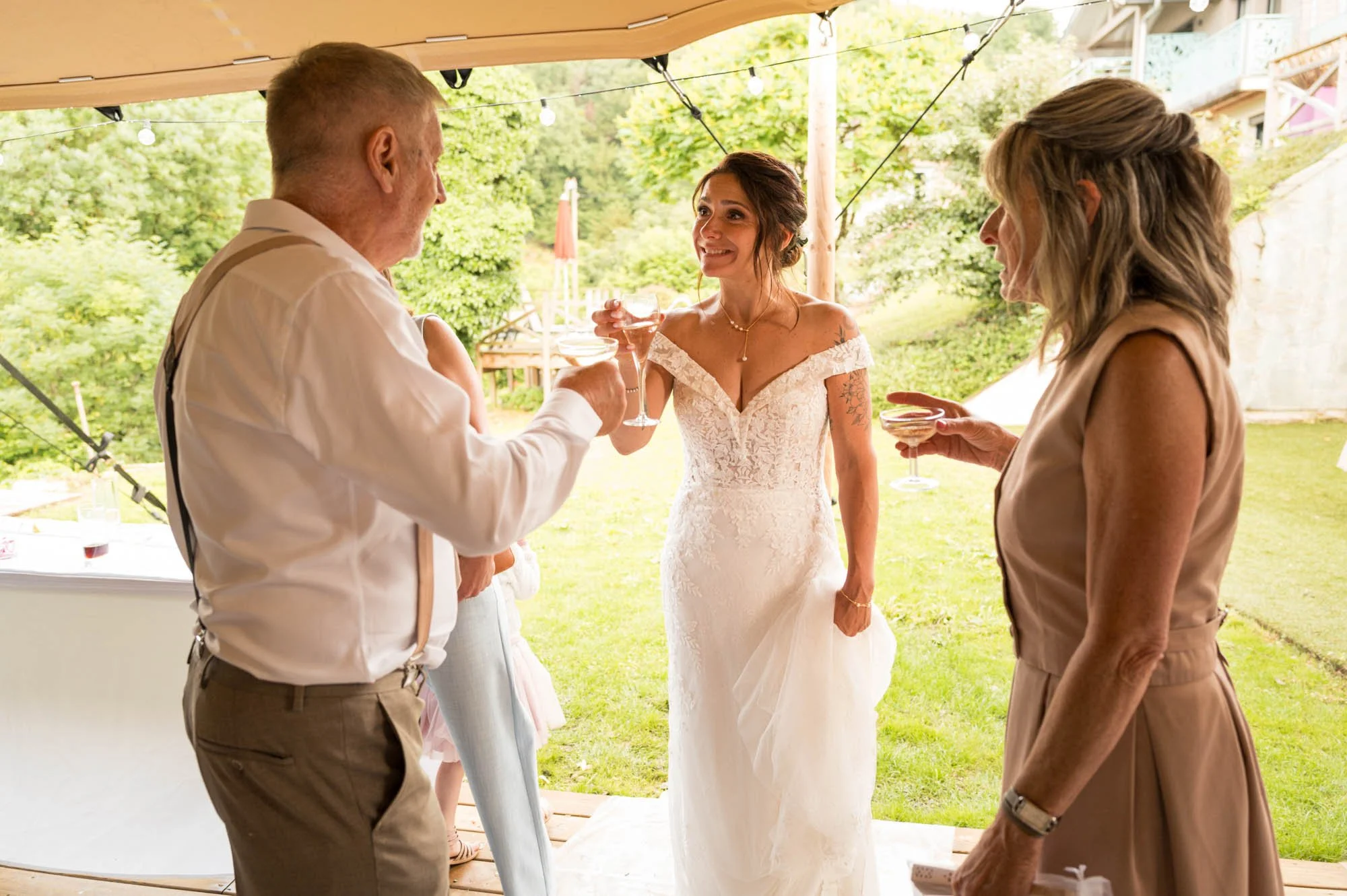 Une femme en robe de mariage souriante, entourée de ses proches, tenant un verre à champagne lors d'une célébration en plein air.