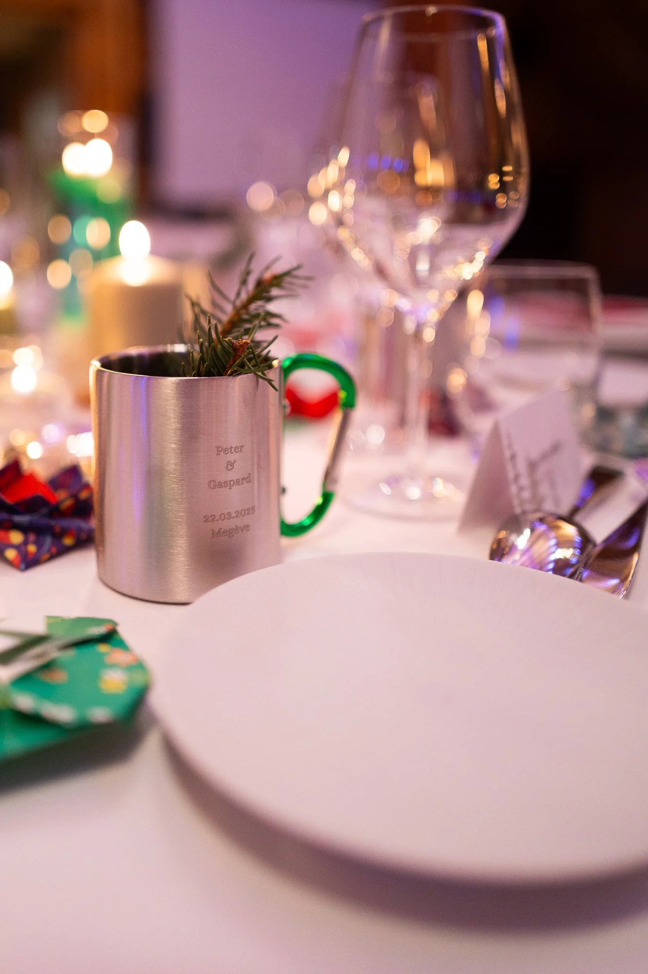 Table de fête avec assiette blanche, verre à vin, tasses en métal, décoration avec une branche de pin, bougies, et une carte de fête.