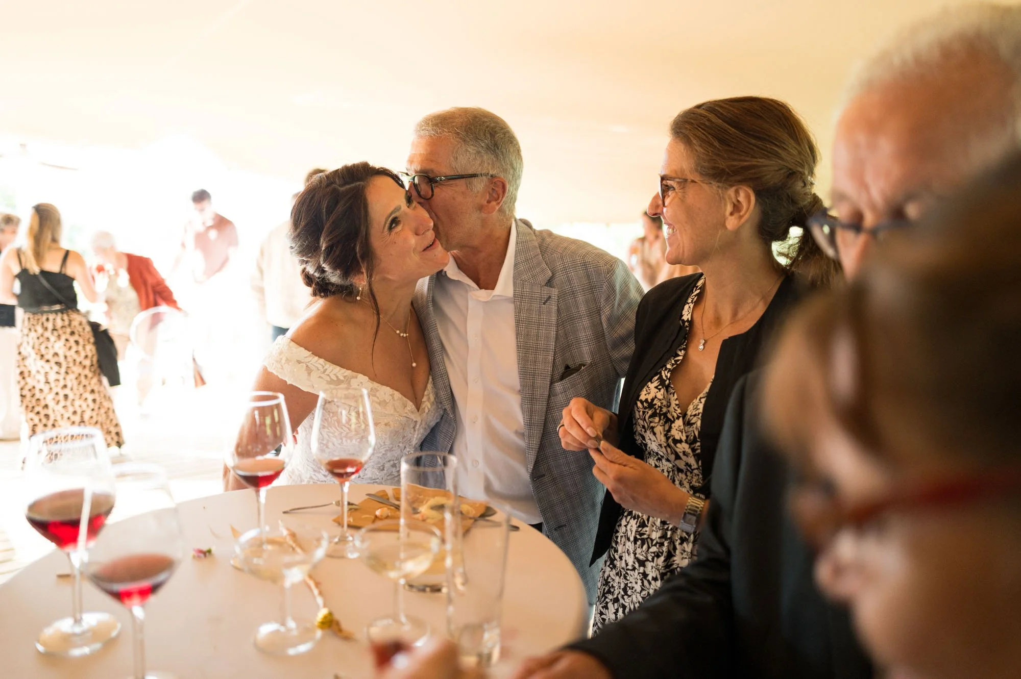 Groupe de personnes lors d'une célébration, avec une femme en robe blanche et un homme en veste à carreaux échangeant un baiser sur la joue, entourés d'autres invités.