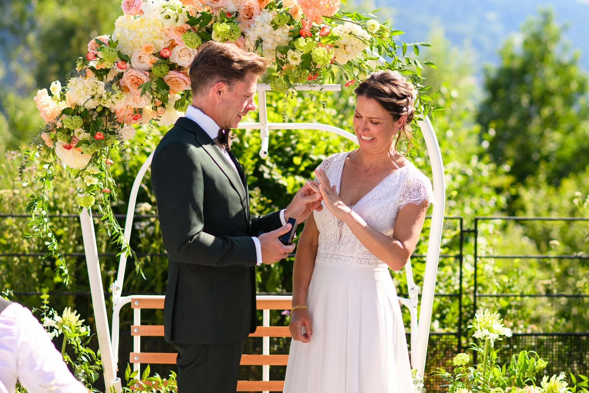 Un couple de mariés échangeant leurs alliances lors de leur mariage en plein air, avec une arche décorée de fleurs roses, blanches et vertes.