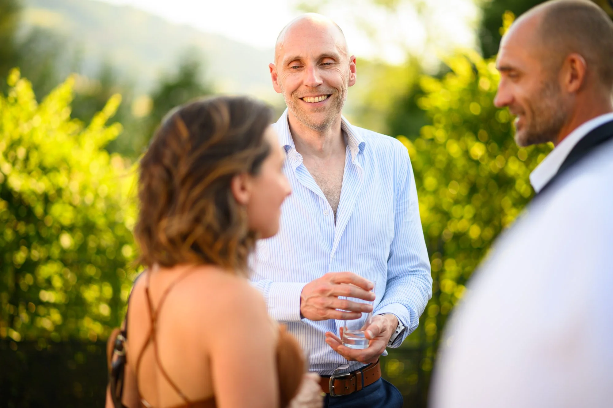 Trois personnes conversent lors d'une réunion en extérieur, sous un soleil brillant, avec un fond de verdure.