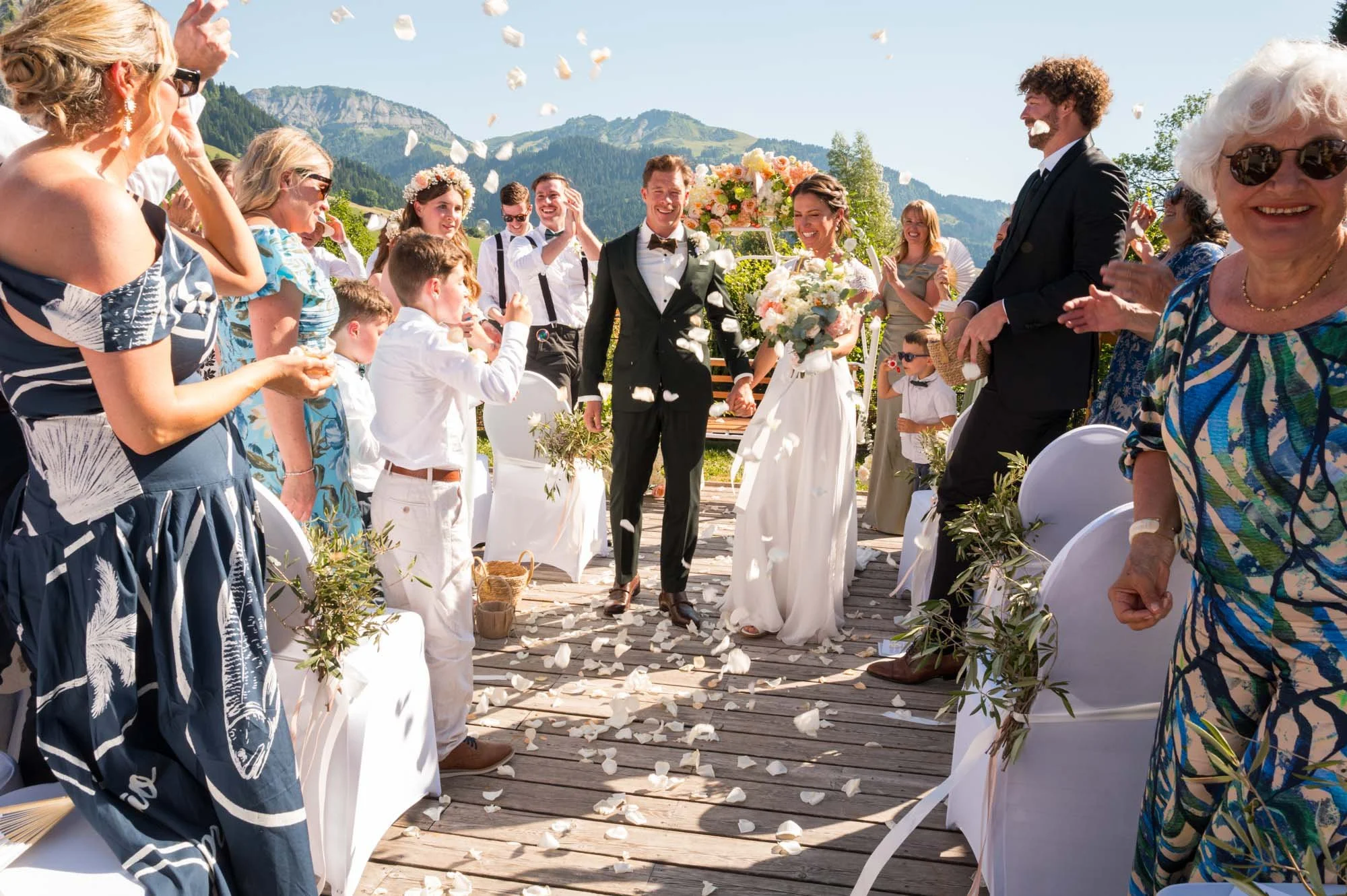 Un mariage en plein air avec un couple de mariés marchant au centre, entouré d'invités applaudissant et lançant des pétales de fleurs, avec des montagnes en arrière-plan.