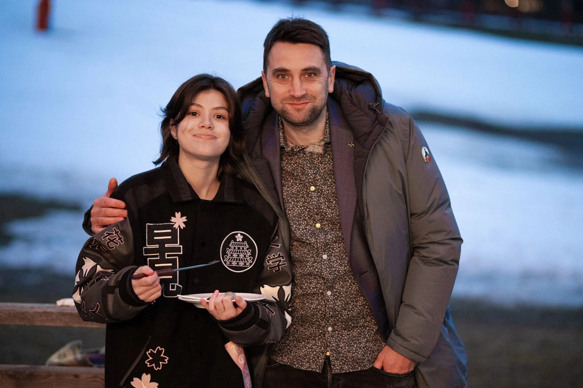 Un homme et une femme posent ensemble près d'une rivière ou d'un lac au coucher du soleil, sourire pour la photo, homme portant une veste grise et femme portant une veste noire avec des motifs.