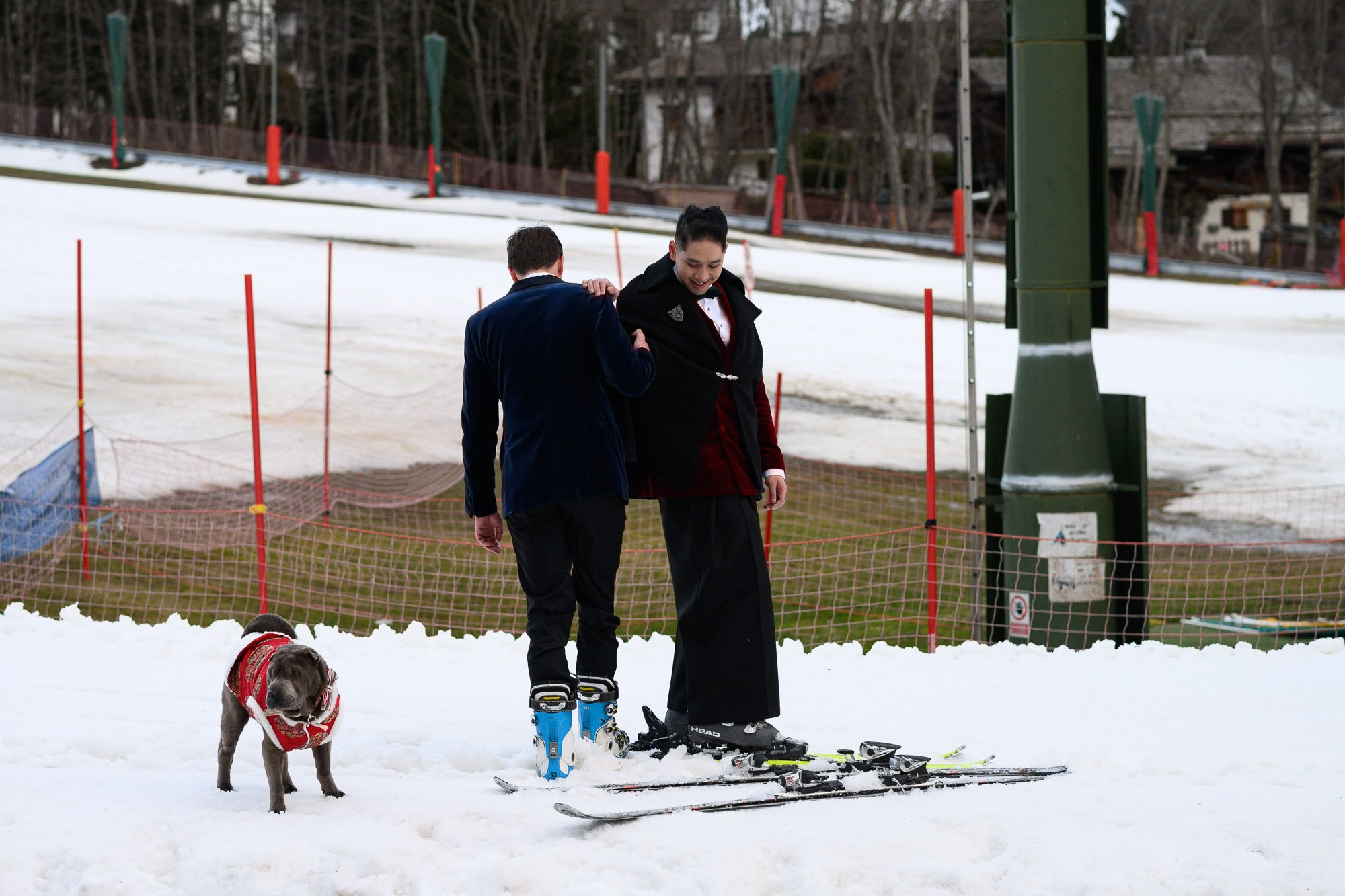Deux personnes habillées de façon élégante, l'une avec des skis et l'autre avec un manteau, se tenant sur la neige, accompagnées d'un chien portant un manteau rouge.