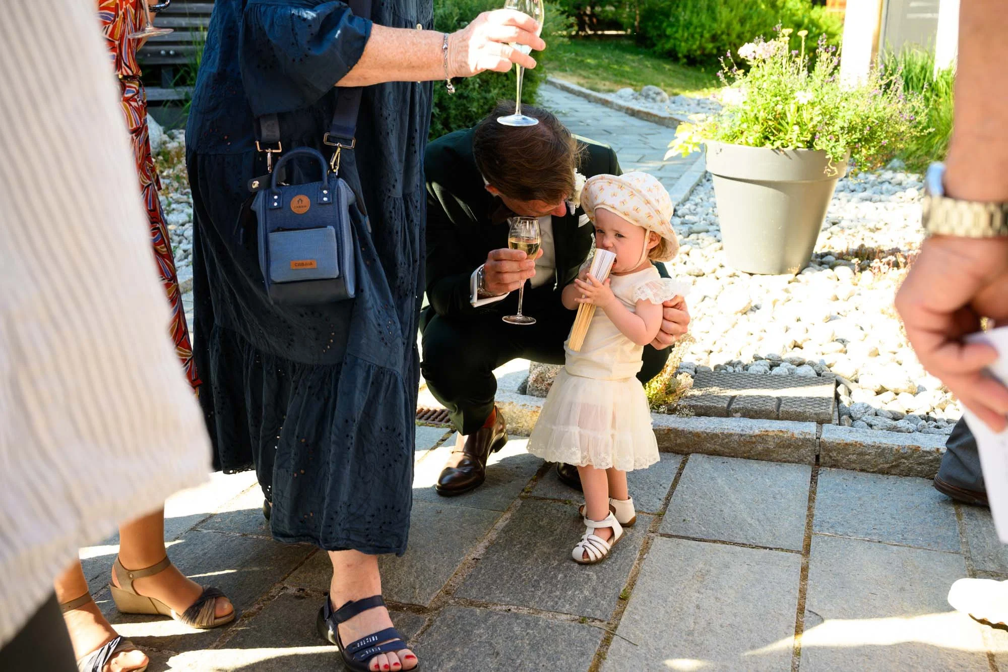 Une petite fille en robe blanche et chapeau à fleurs, tenant un rouleau et une baguette, est entourée de plusieurs adultes lors d'une fête extérieure. Un homme à genoux lui parle, et plusieurs personnes tiennent des verres de champagne.