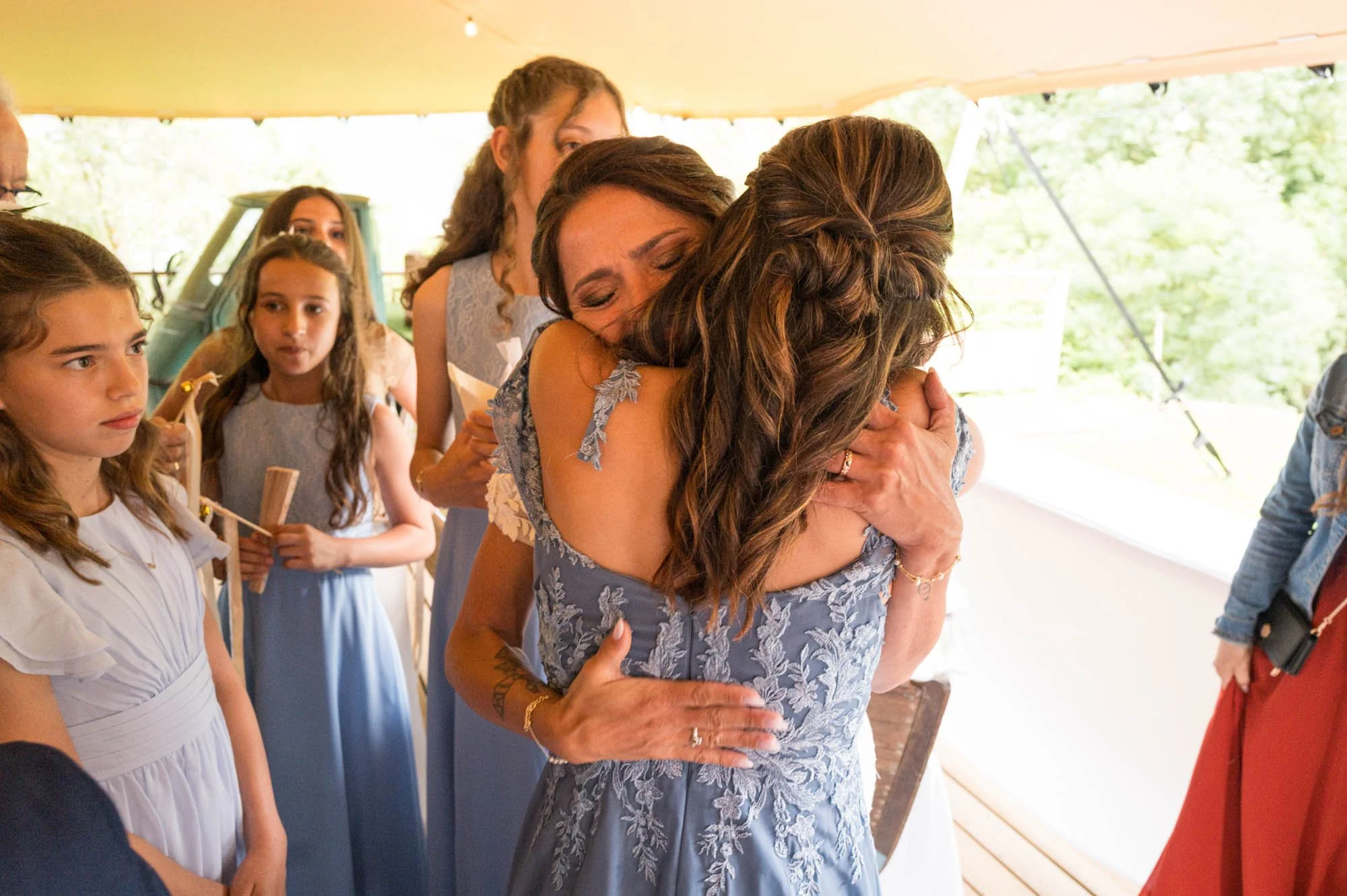 Deux femmes s'embrassent dans un câlin émouvant lors d'un mariage, entourées de jeunes filles en robes de cérémonie.