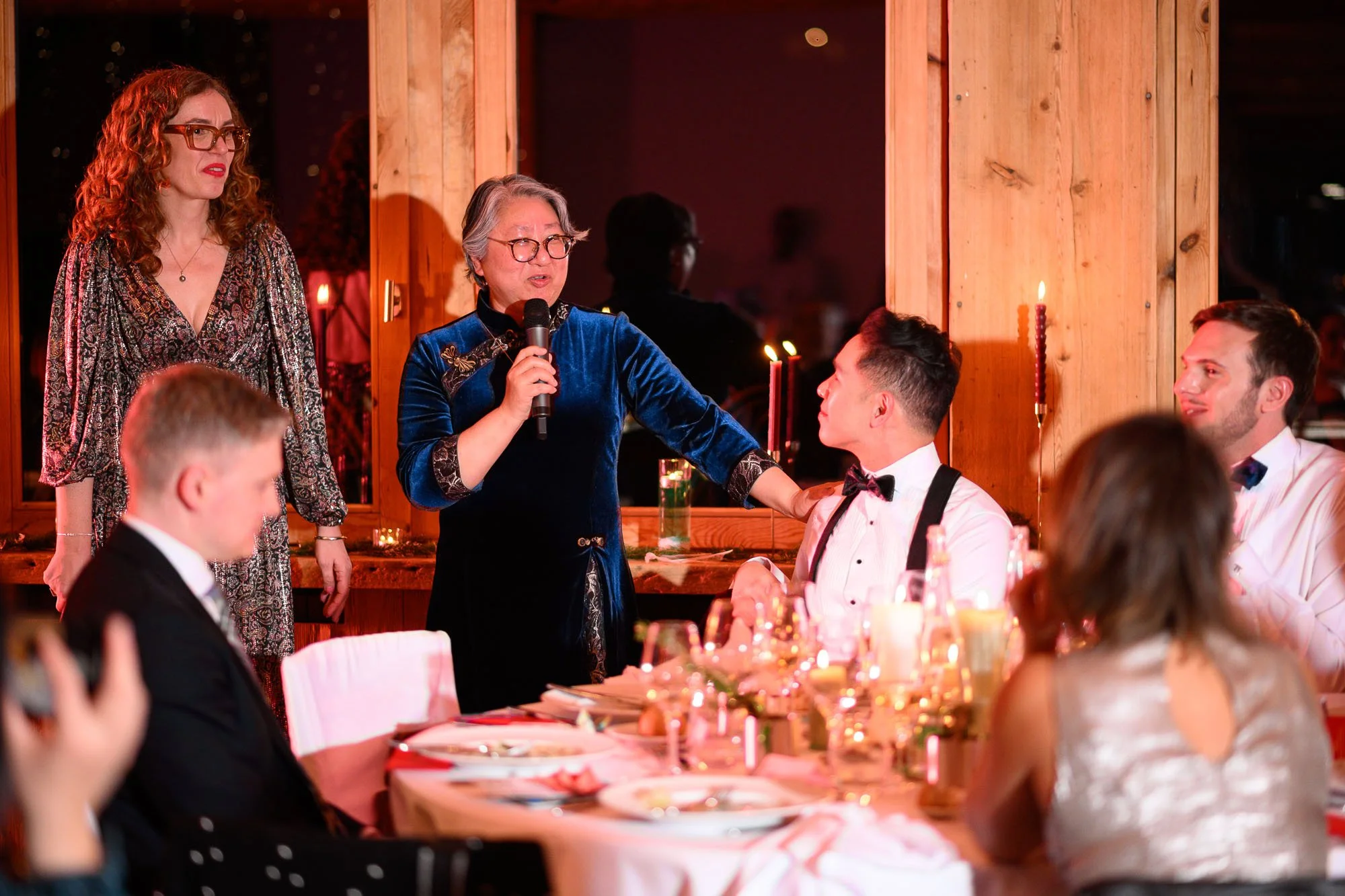 Une scène de discours lors d'une fête ou d'un dîner formel, avec une femme âgée parlant dans un microphone, entourée de personnes assises à une table décorée
