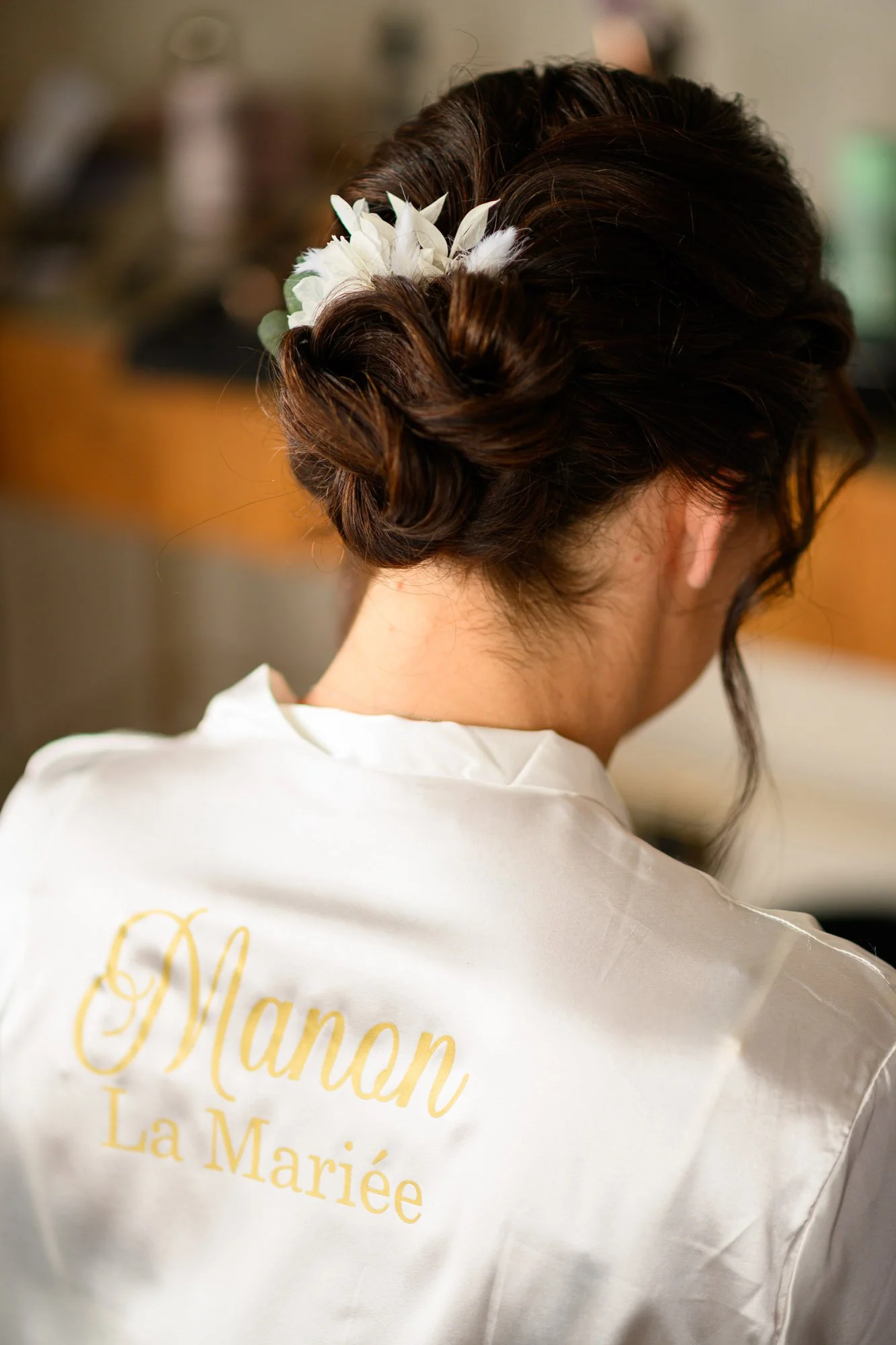 Une femme avec une coiffure élaborée porte une robe en satin blanc avec l'inscription "Manon La Mariée" en doré dans le dos.