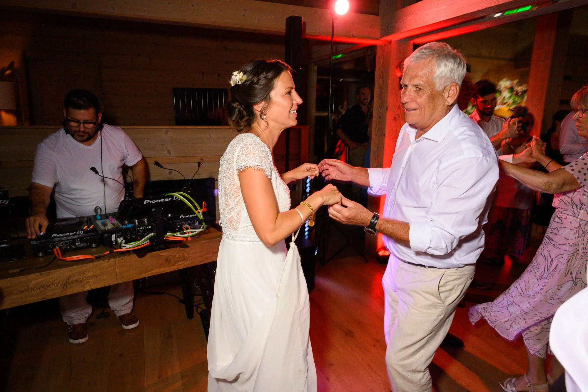 Une femme en robe blanche danse avec un homme âgé lors d'une fête, avec un DJ à l'arrière-plan et d'autres personnes qui dansent en arrière-plan.