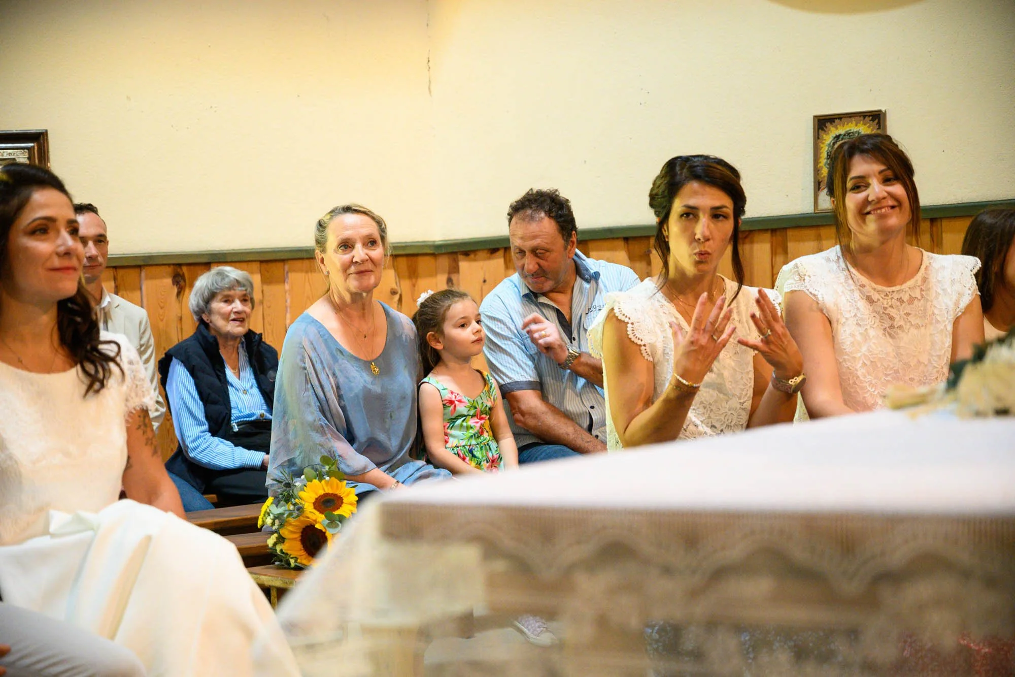Groupe de personnes assises dans une cérémonie, probablement un mariage, avec un décor en bois et des peintures sur le mur. Parmi eux, une femme en robe blanche, une jeune fille en robe florale, et plusieurs femmes en vêtements légers.