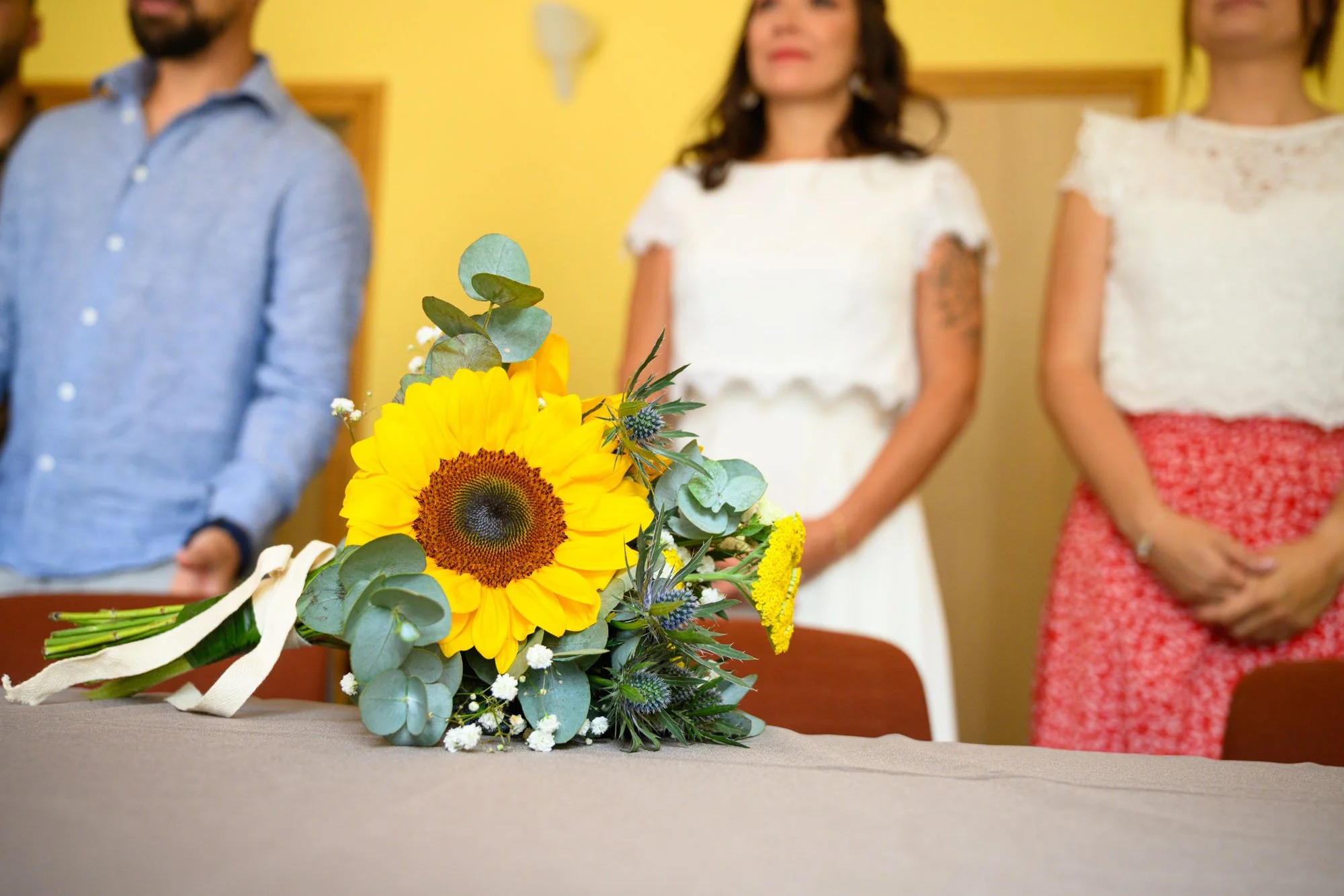 Un bouquet de fleurs avec un tournesol en première plan, posé sur une table. En arrière-plan, trois personnes se tiennent debout, ce sont deux femmes et un homme, dans une pièce aux murs jaunes.