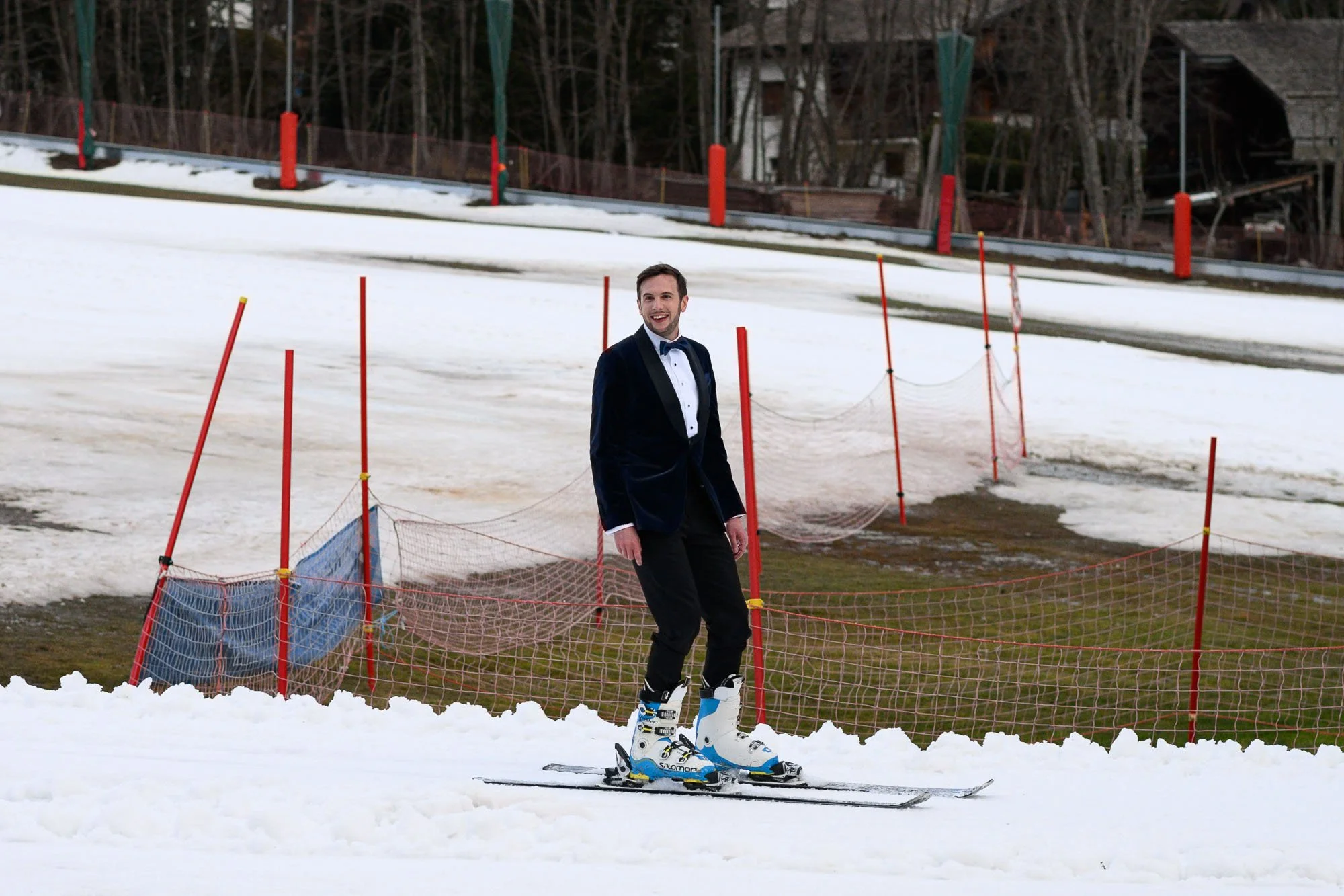 Un homme en costume de smoking sur des skis dans un environnement de ski, avec un peu de neige et des barrières de sécurité en arrière-plan.