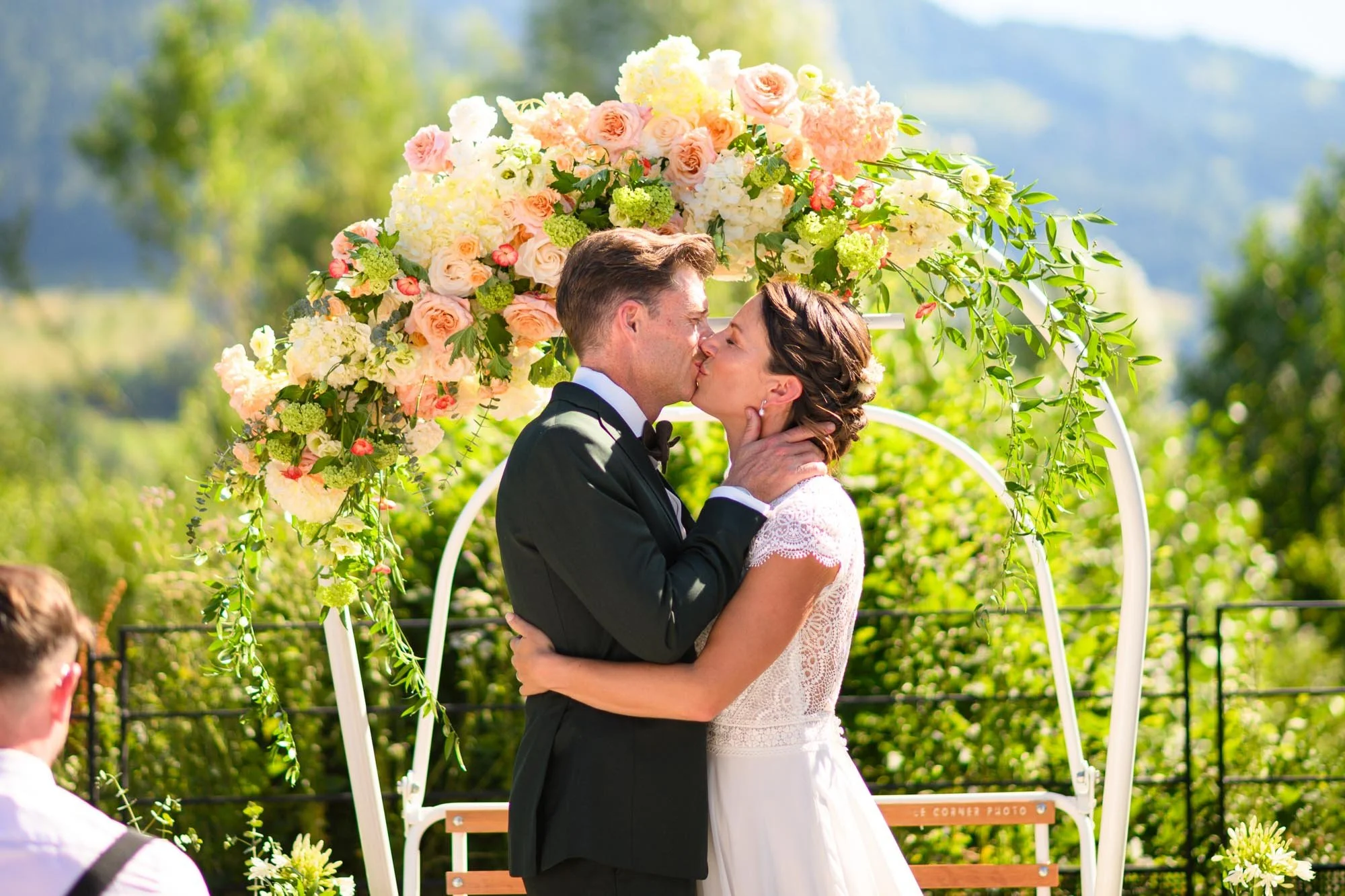 Un couple de mariés s'embrasse lors de leur mariage à l'extérieur, devant un décor floral avec un ciel clair et un paysage verdoyant en arrière-plan.