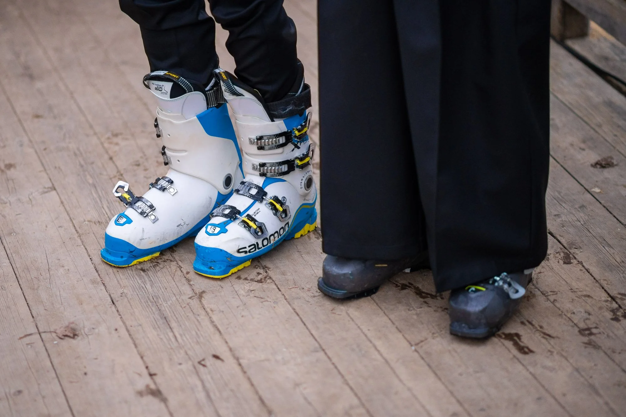 Chaussures de ski alpins blanches et bleues avec des boucles en métal et un logo Salomon, situées sur un plancher en bois à côté de bottes de ski sombres.
