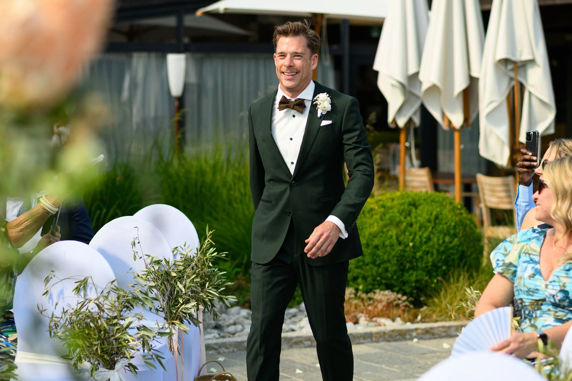 Un homme en smoking noir avec une boutonnière blanche à une cérémonie de mariage en plein air, souriant, entouré de spectateurs qui prennent des photos.