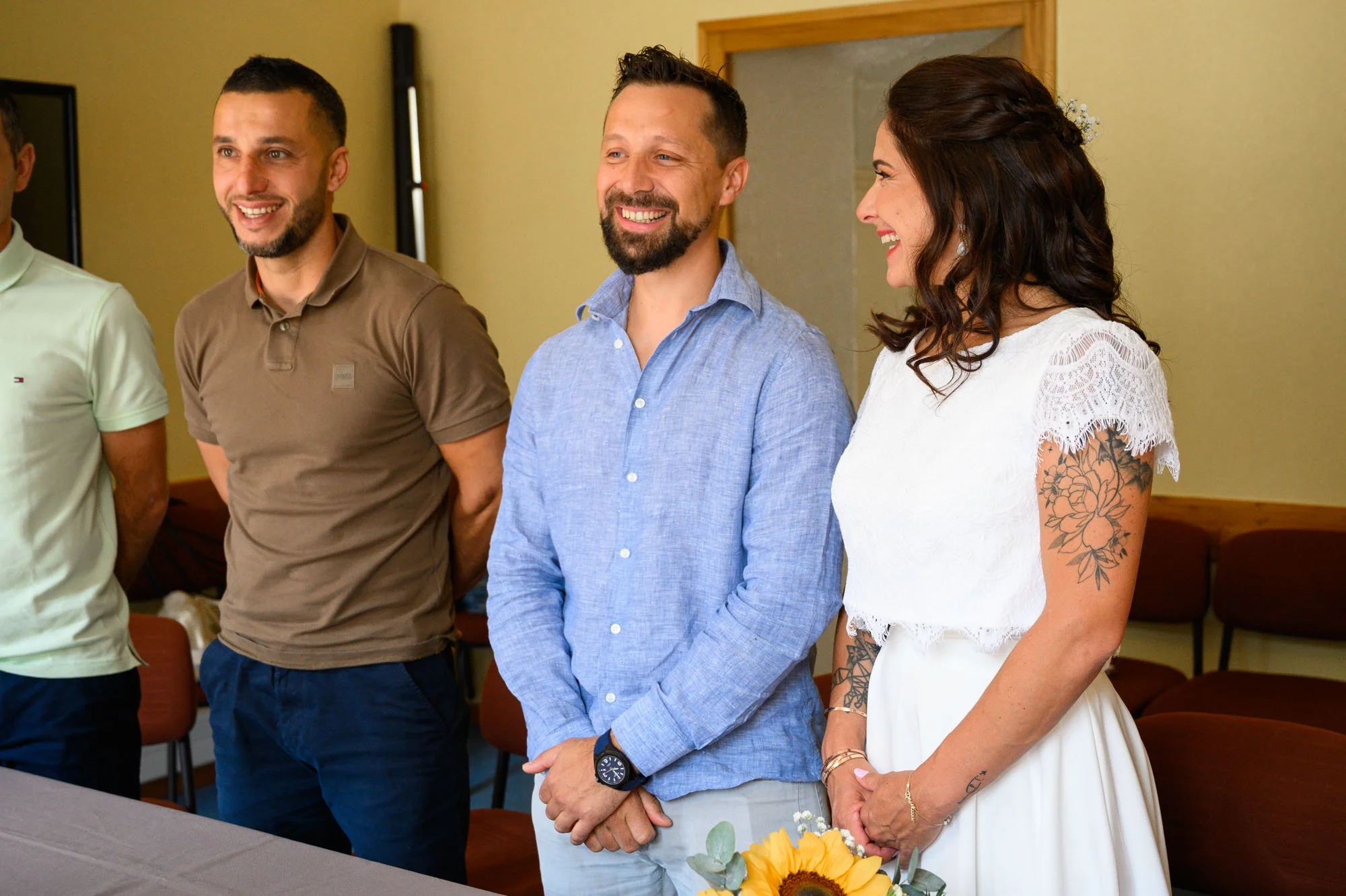 Trois personnes debout en ligne dans une pièce, souriant, lors d'une occasion sociale ou événement formel, avec une femme tenant un bouquet de tournesols.