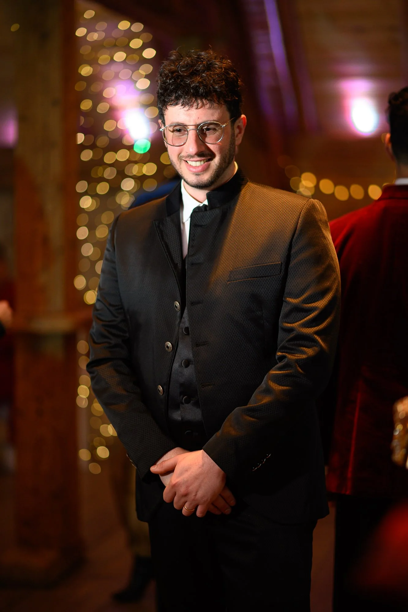 Un homme souriant, portant un costume noir élégant avec des boutons, debout dans un lieu décoré avec des lumières et des guirlandes, suggérant une ambiance festive ou un événement spécial.