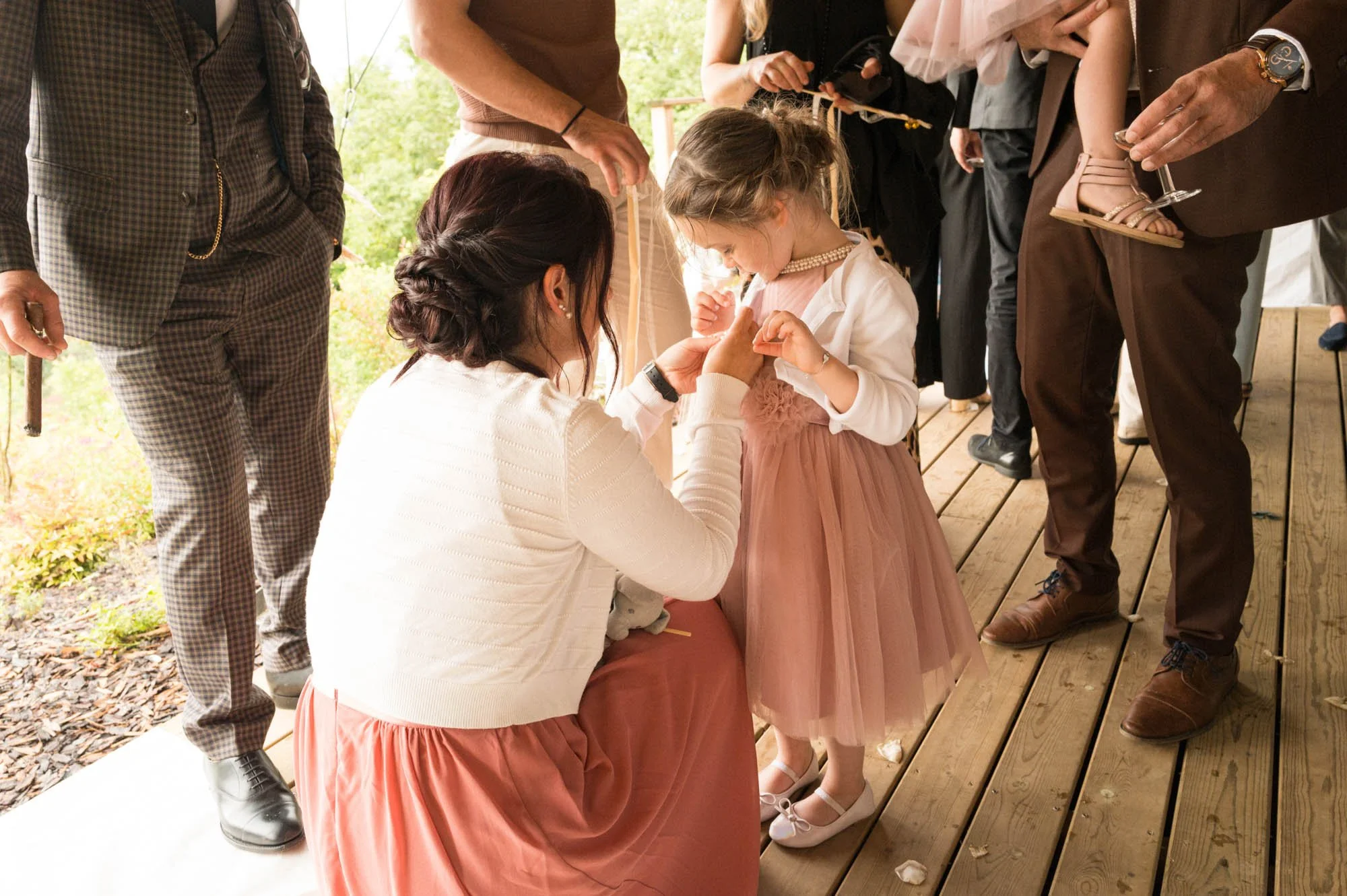 Une femme agenouillée attache la boutonnière d'une petite fille lors d'un mariage, entourée de plusieurs adultes, sur un pont en bois à l'extérieur.