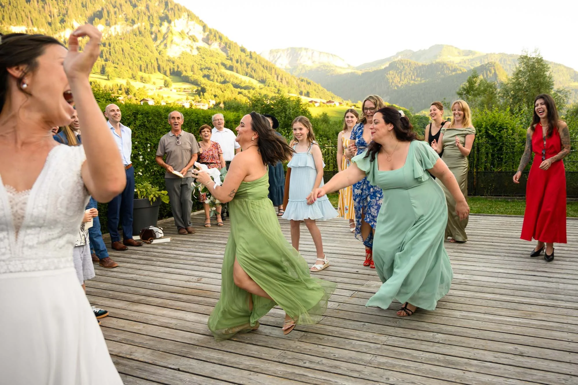Groupe de personnes dansant et riant lors d'une fête en plein air sur une terrasse en bois avec un paysage montagneux en arrière-plan.