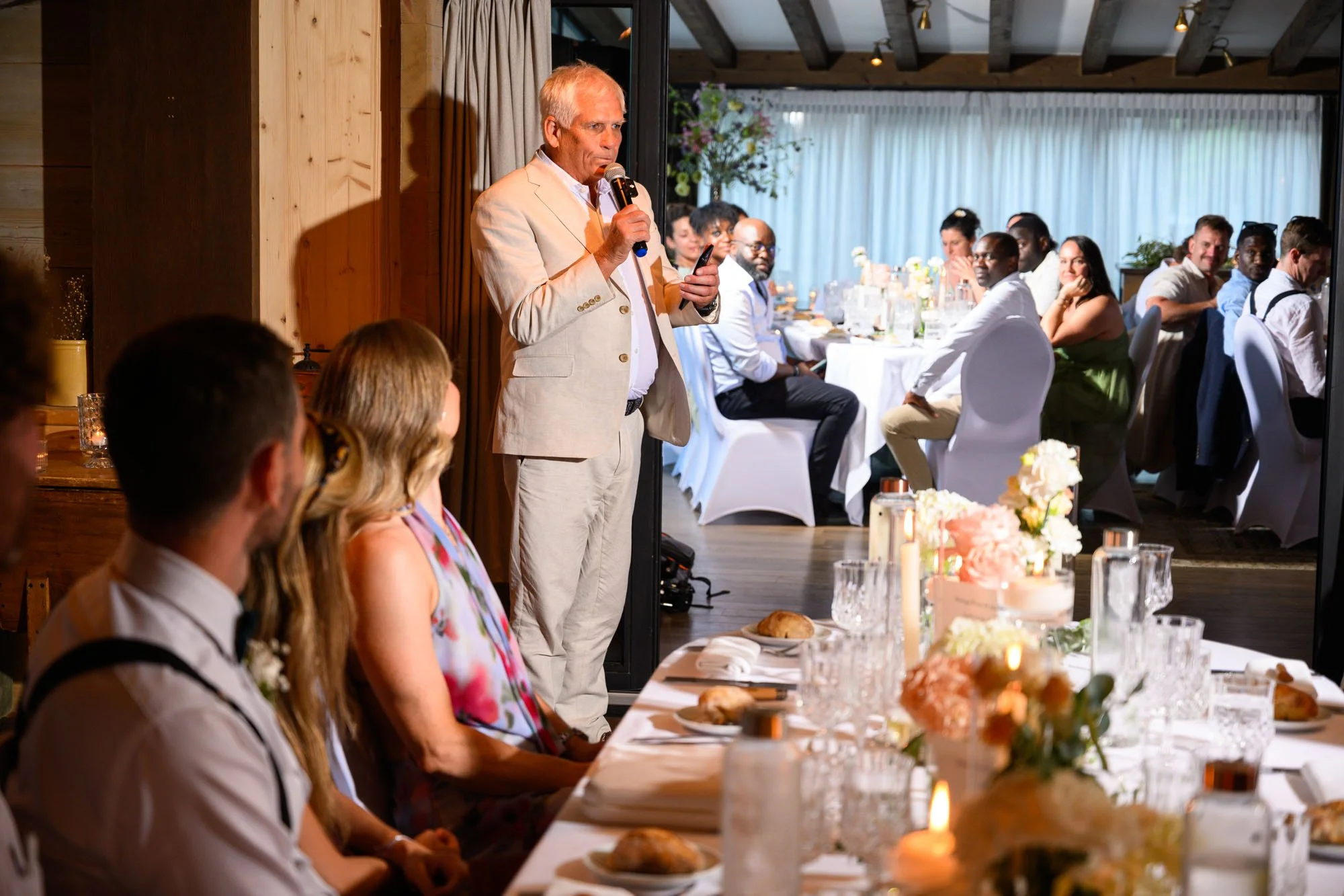 Un homme en costume clair parle avec un microphone lors d'un discours à un événement ou un mariage, avec un groupe d'invités assis à une table décorée de fleurs et de bougies en arrière-plan.