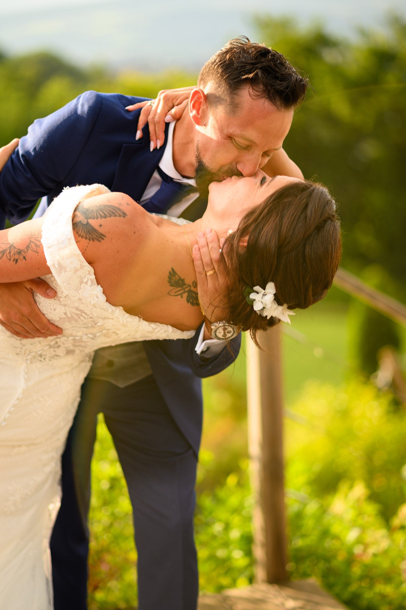 Un couple en habit de mariage partage un baiser lors de leur mariage en plein air, avec un fond de nature verdoyante.