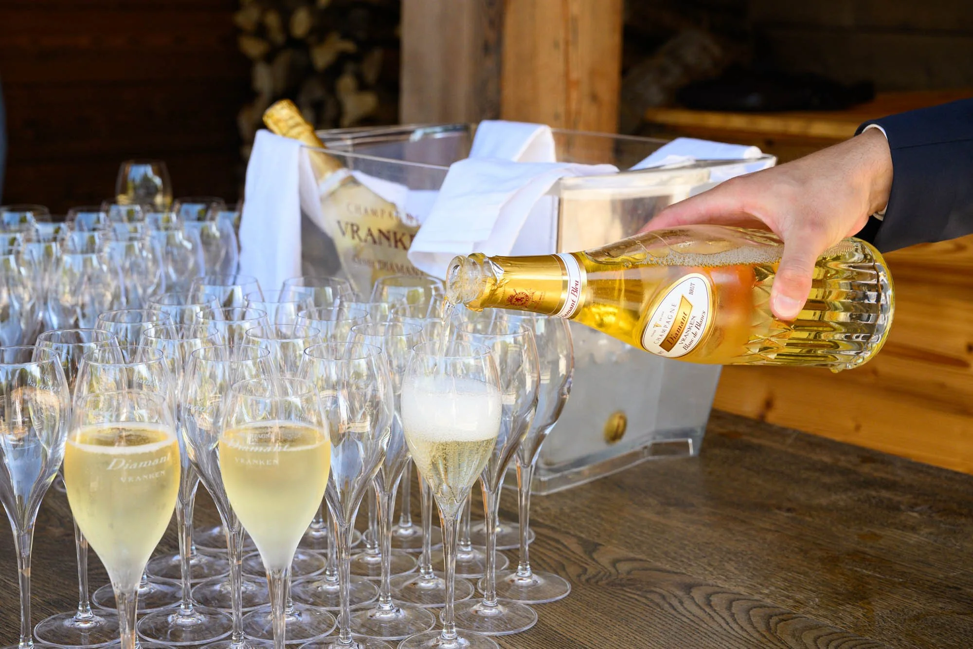 Une personne verse du champagne dans des coupes en cristal, avec un seau à champagne rempli de bouteilles de Champagne Vrancken en arrière-plan.