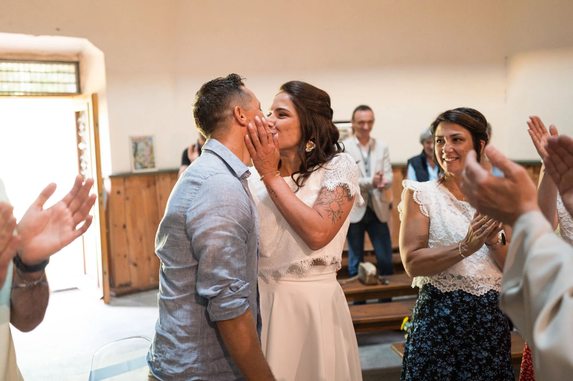Deux personnes s'embrassent lors d'une cérémonie, entourées de personnes applaudissant dans une salle lumineuse.