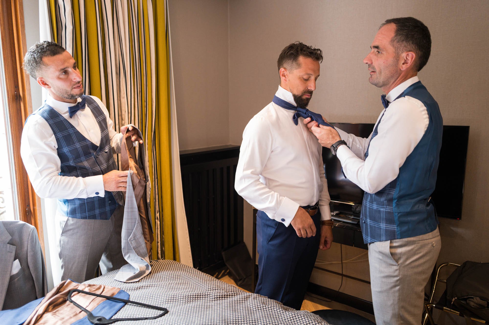 Trois hommes habillés en costumes en train de se préparer pour un événement, avec deux d'entre eux essayant des nœuds papillon.