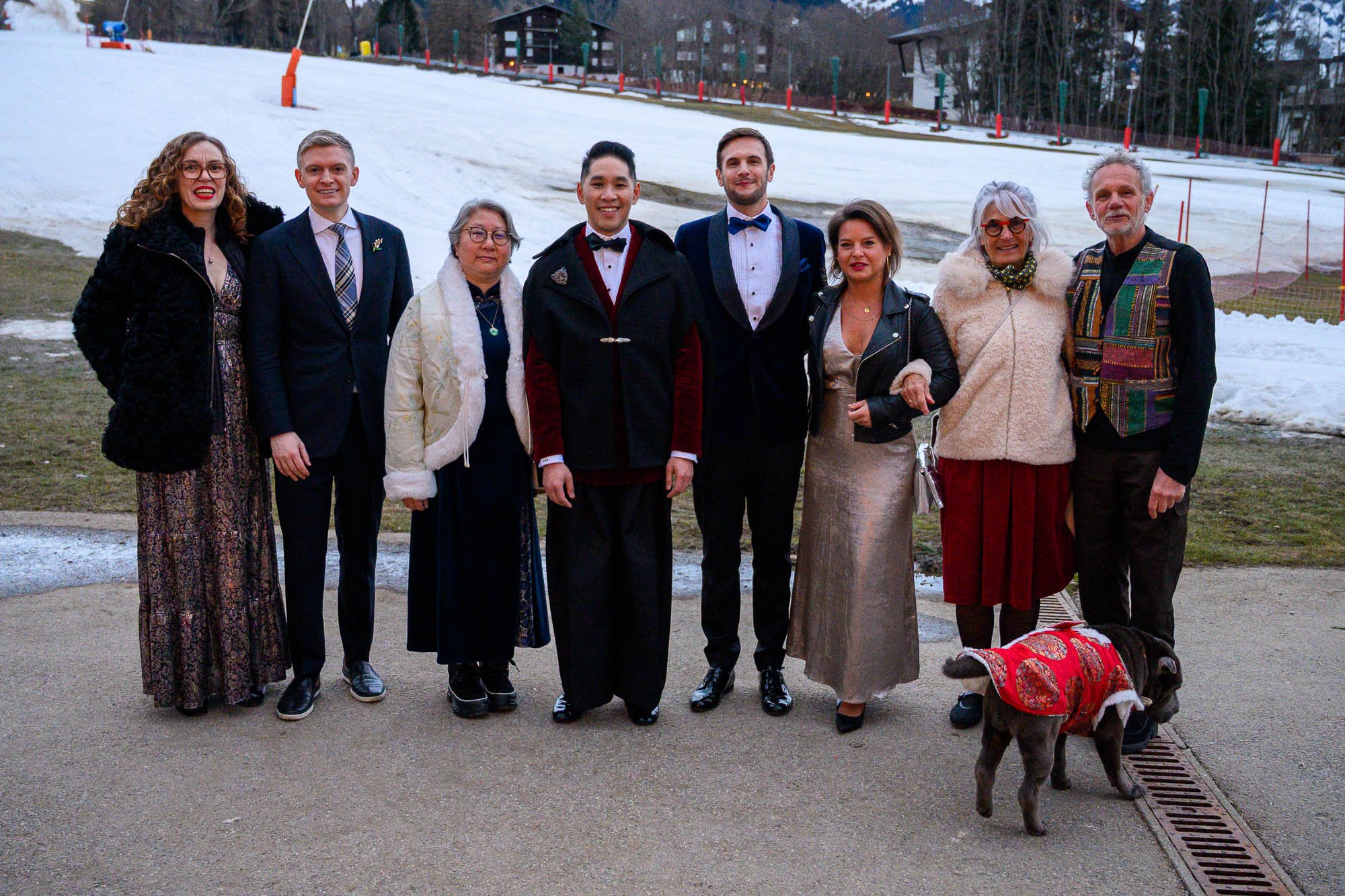 Groupe de neuf personnes habillées de façon élégante, posant devant un paysage enneigé avec des bâtiments et un terrain de ski en arrière-plan. Présence d'un chien portant un manteau rouge.