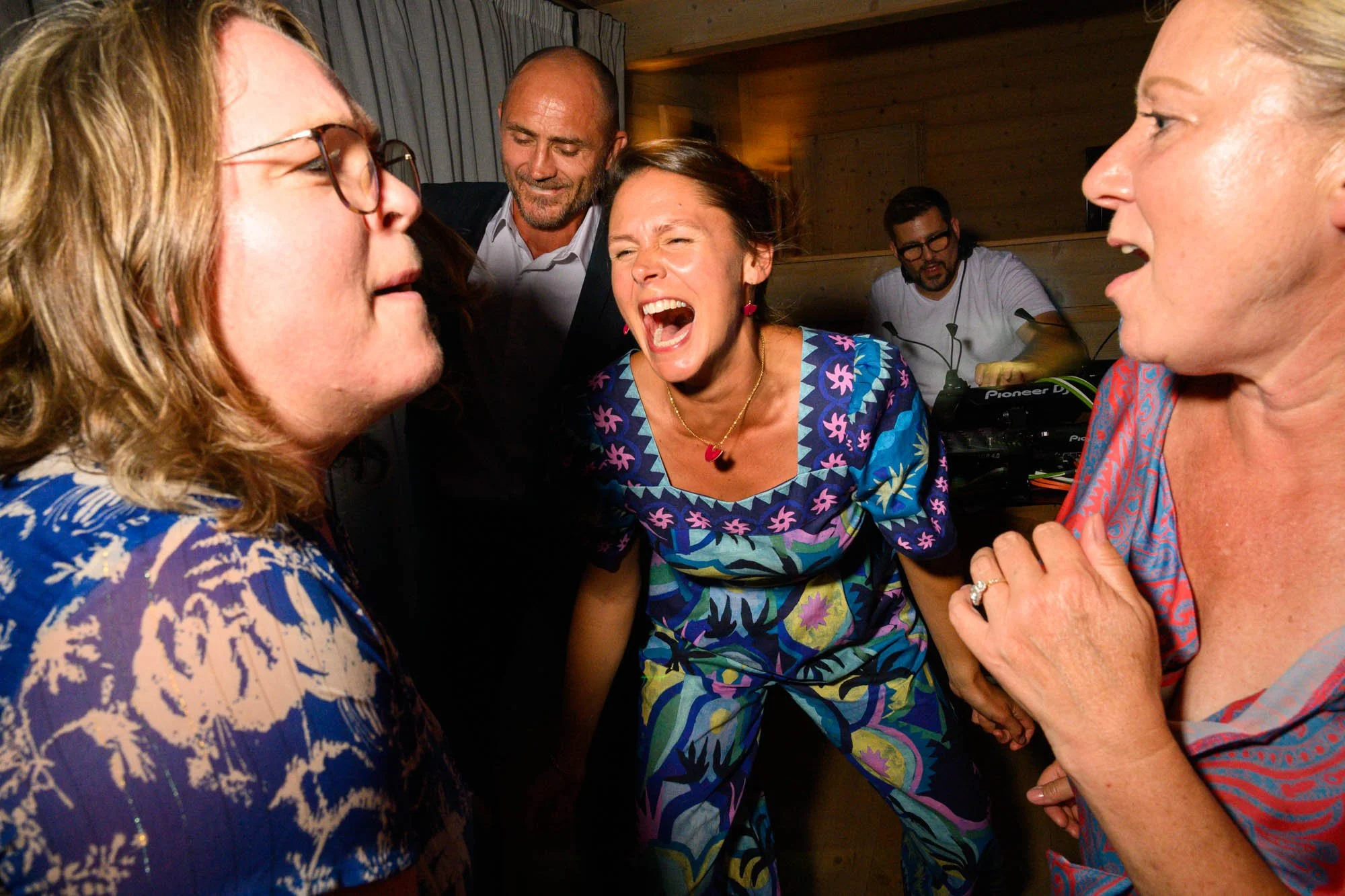 Groupe de personnes qui chantent ou crient lors d'une fête, dans une pièce en bois, avec un DJ à l'arrière-plan.