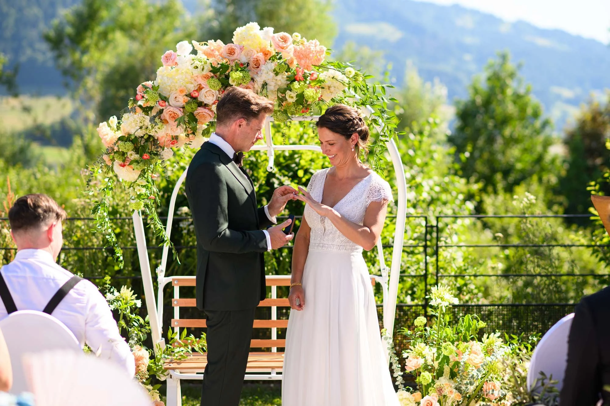 Un couple de mariés échange leurs vœux lors d'une cérémonie en plein air avec un décor floral. La mariée porte une robe blanche et le marié un costume noir.