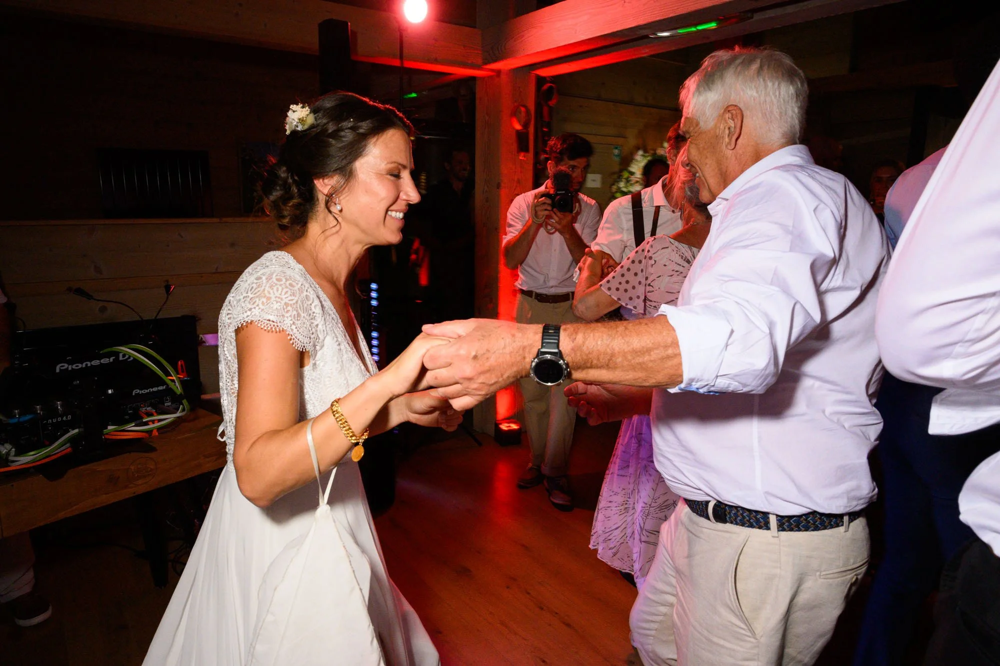 Une femme en robe blanche et un homme âgé dans une chemise blanche dansant et se tenant la main lors d'une célébration, probablement un mariage, dans une ambiance chaleureuse.