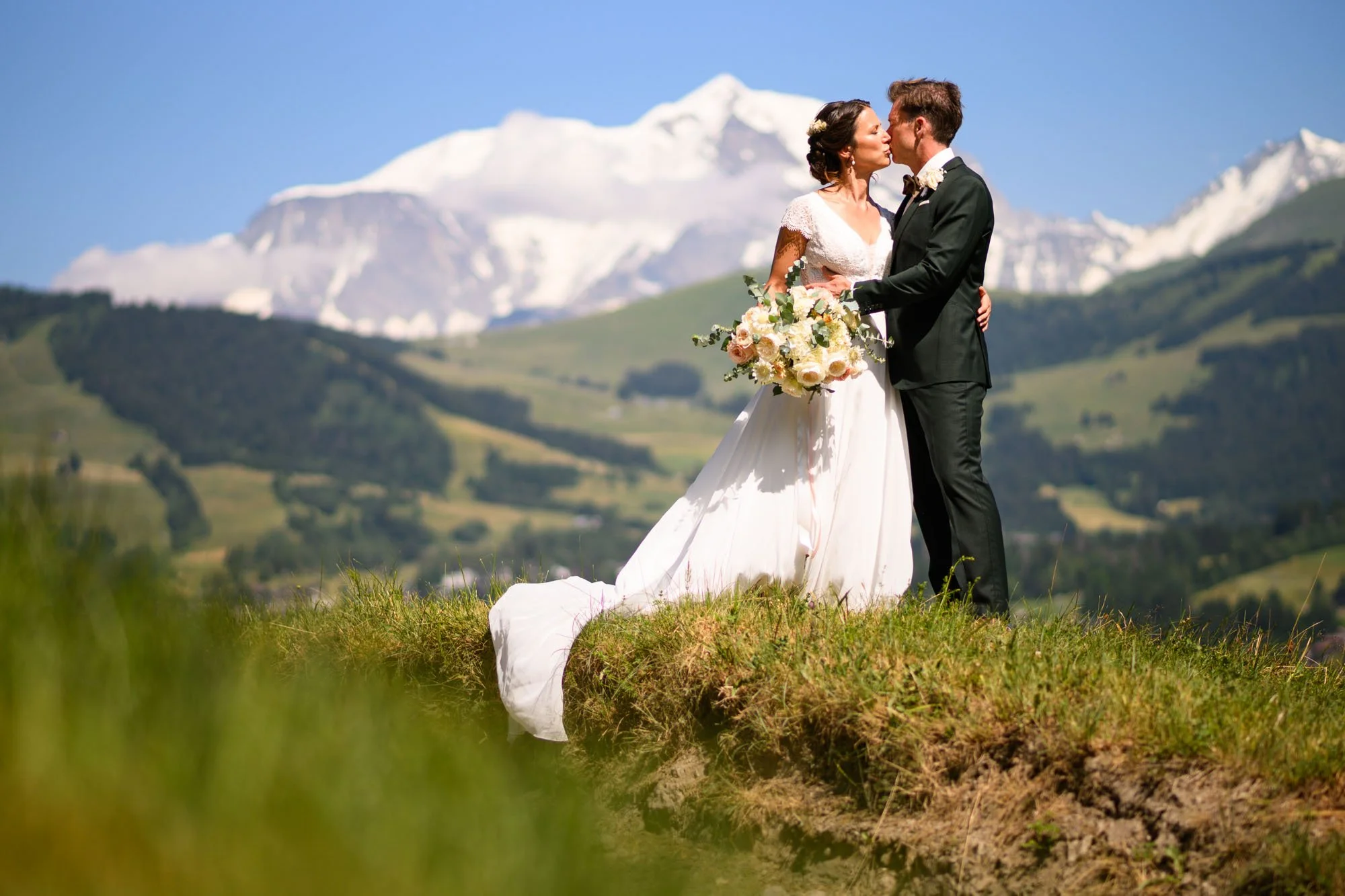 Un couple de mariés s'embrassant lors d'une séance photo en plein air avec un fond de montagnes enneigées et un ciel bleu.
