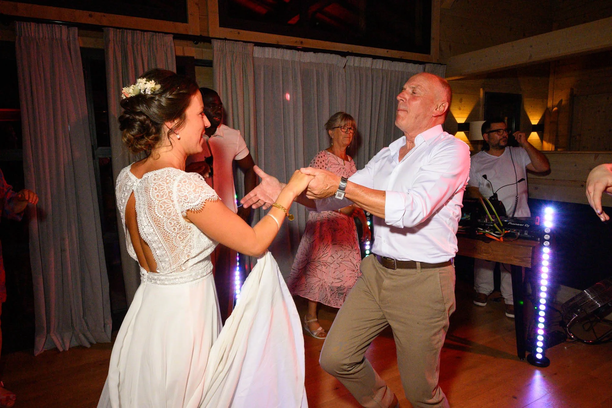 Un homme et une femme dansent ensemble lors d'une fête, d'autres personnes regardent en arrière-plan, ambiance chaleureuse dans une pièce en bois.
