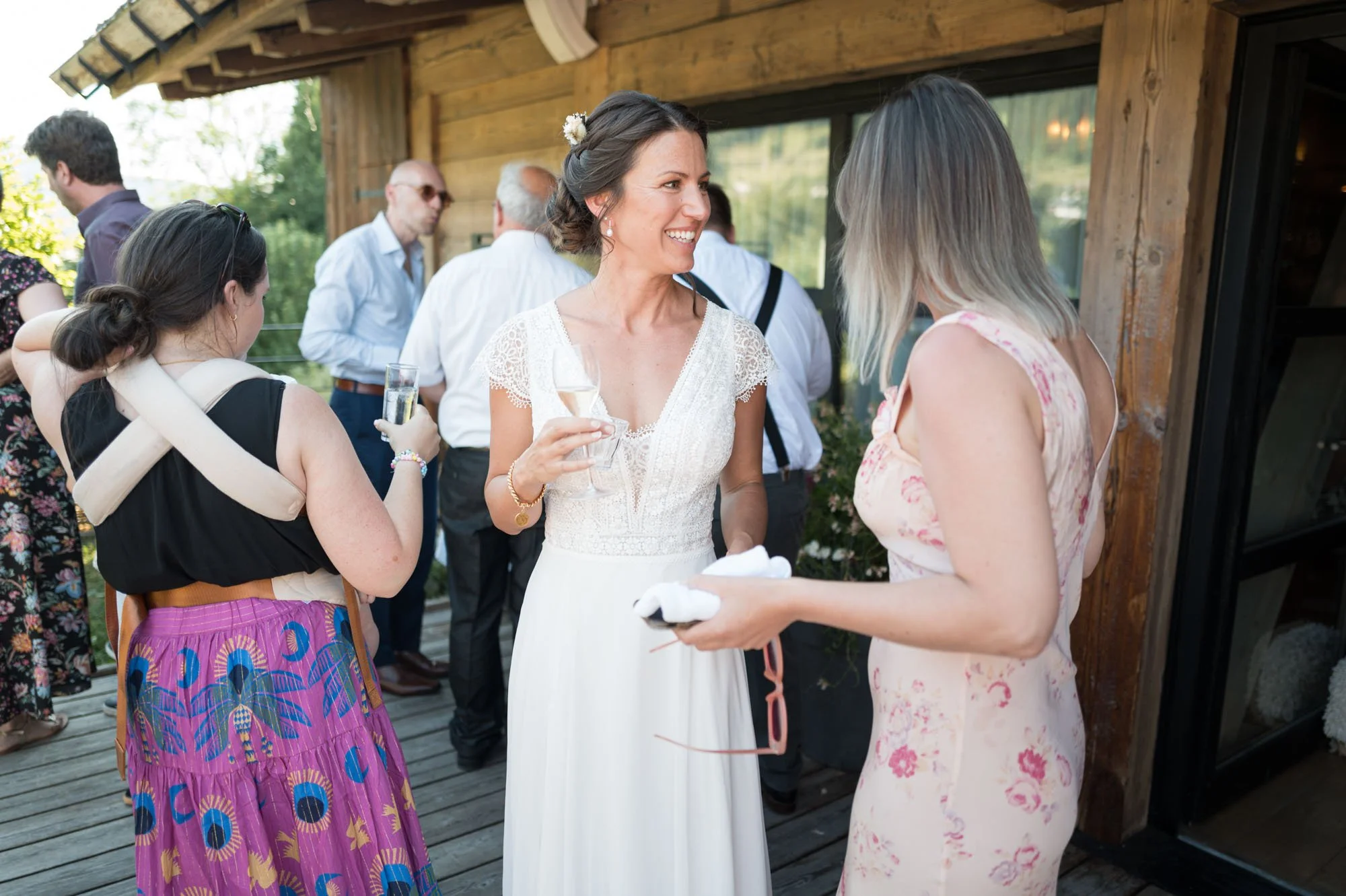 Deux femmes discutent lors d'une célébration en plein air, une porte-claire, en robe blanche, tenant un verre de champagne, souriante, et une autre en robe rose pâle, avec des lunettes, tenant un vêtement ou un accessoire, entourées d'autres invités 