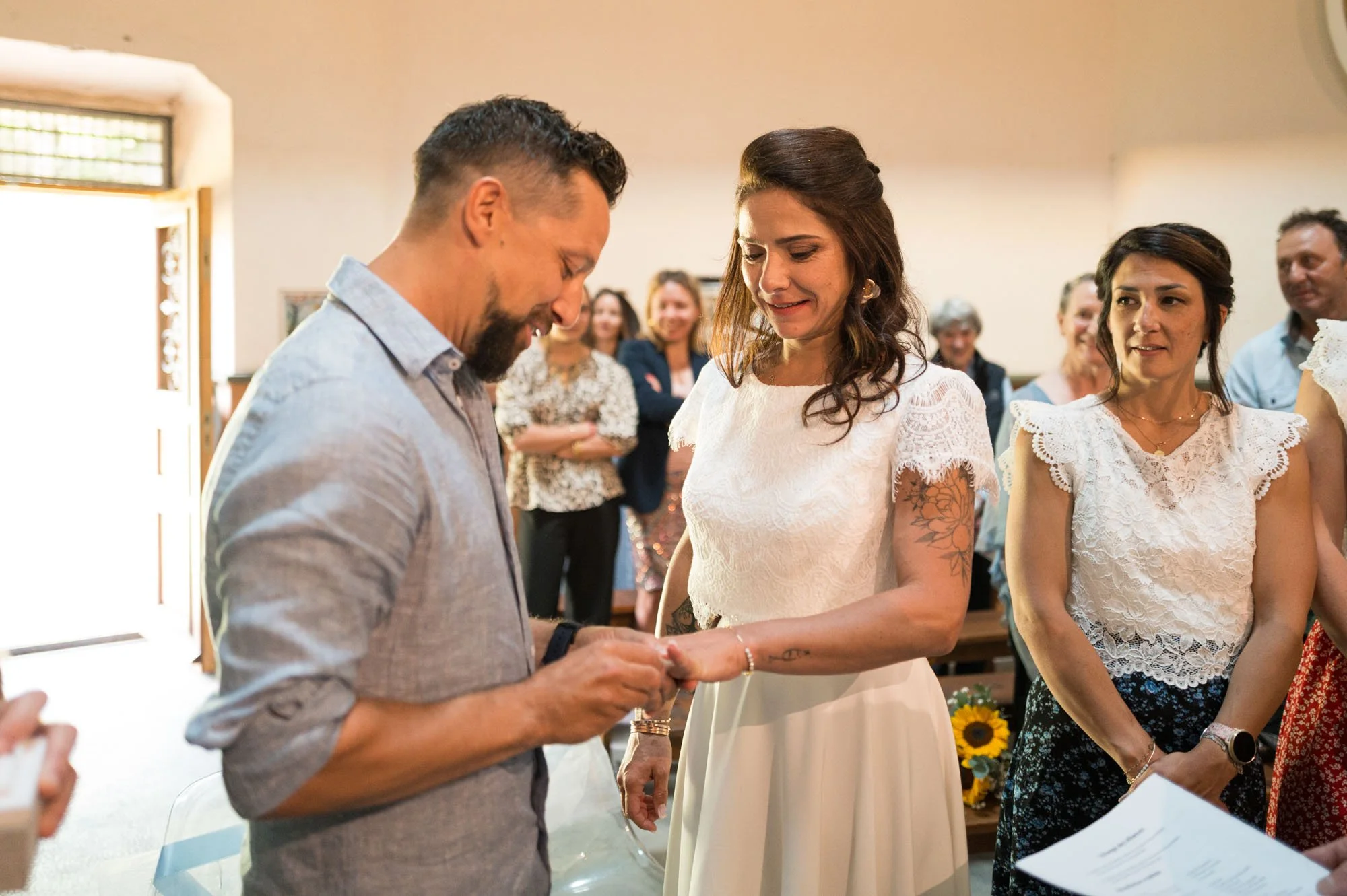 Une femme en robe blanche de mariage et un homme en chemise grise échangent des vœux lors d'une cérémonie de mariage, entourés de famille et amis.