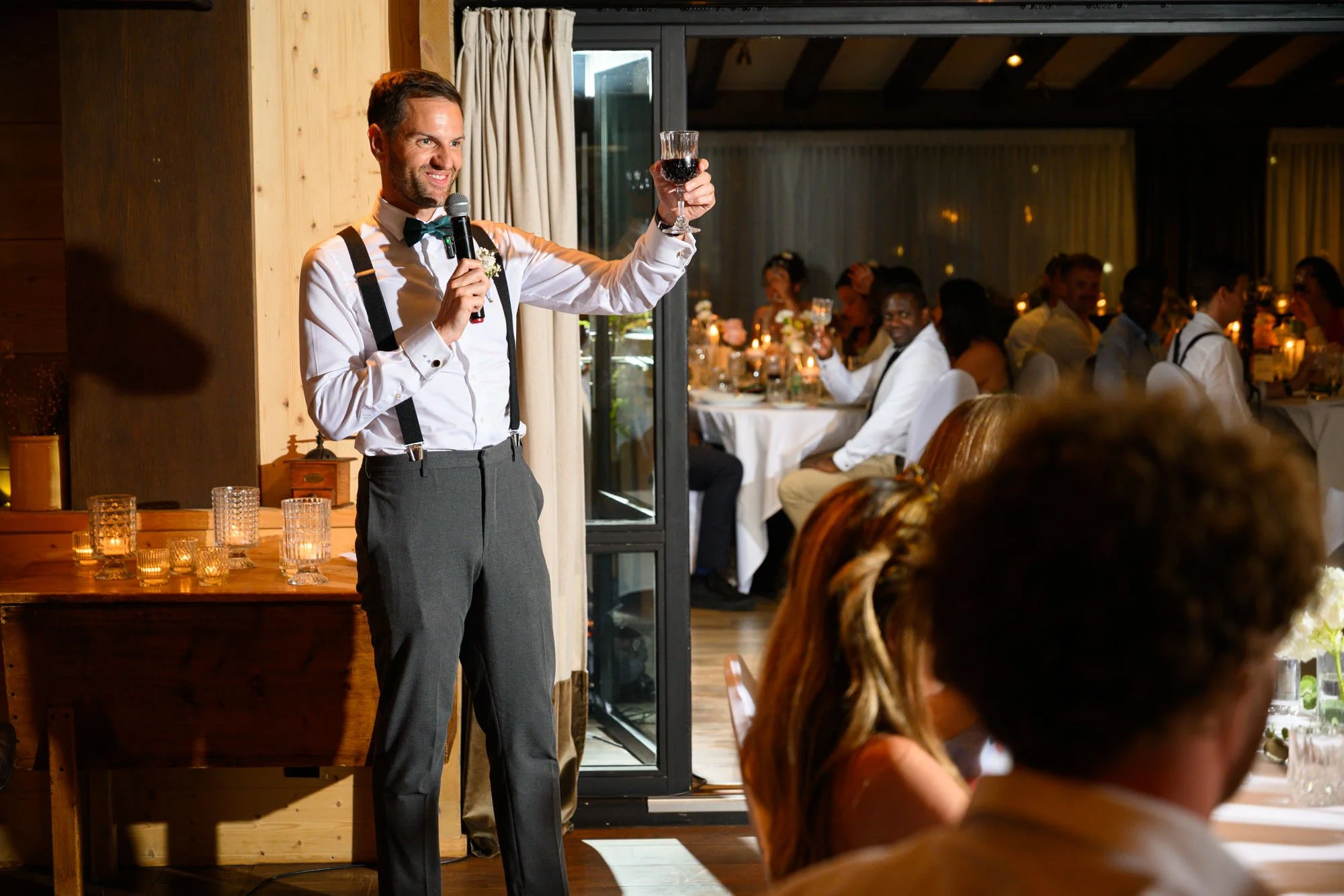 Un homme en tenue de soirée, tenant un micro et un verre de vin rouge, faisant un toast lors d'un événement. Autour, des invités assis à des tables décorées, célébrant dans une ambiance chaleureuse.