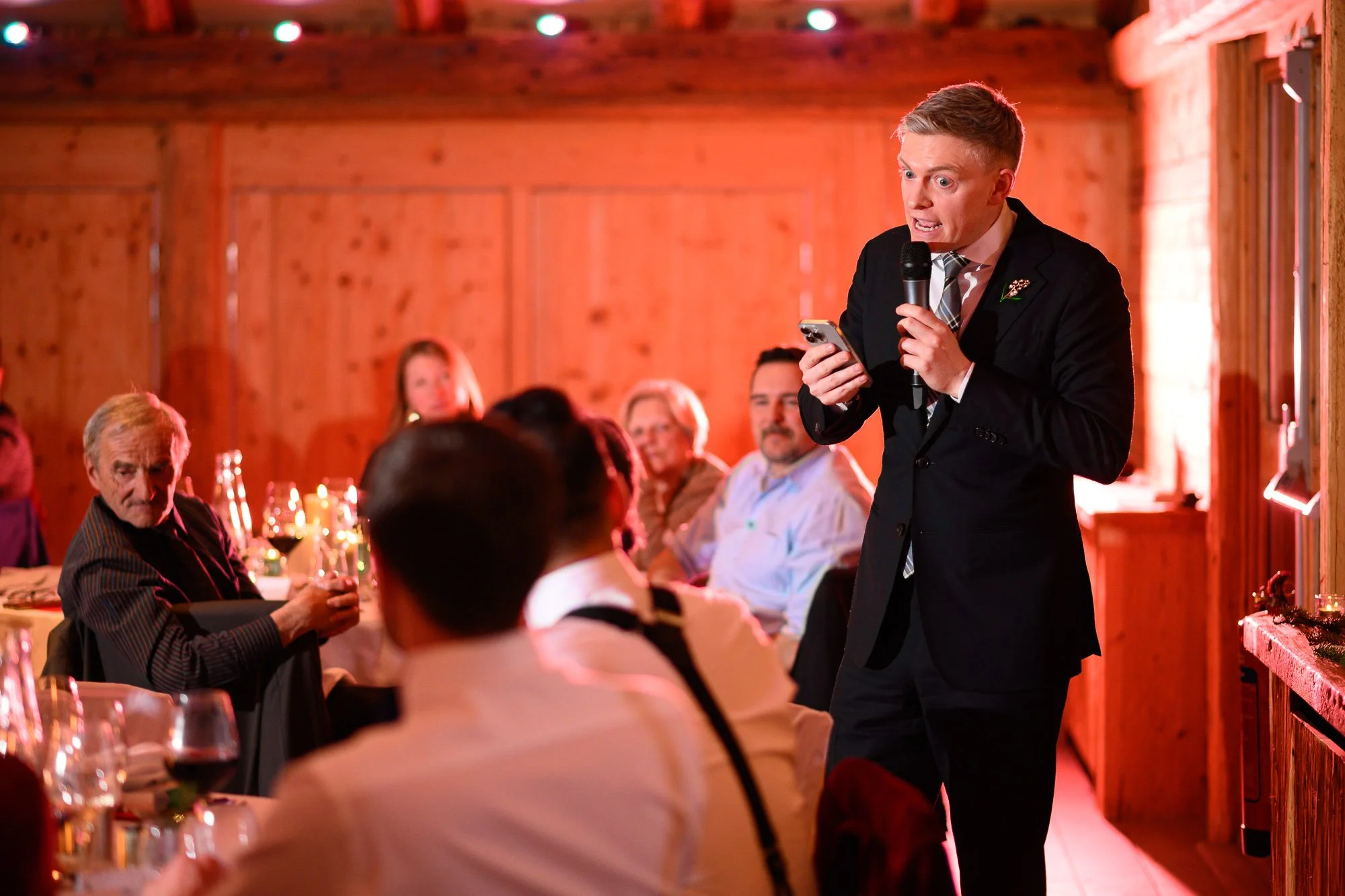 Un homme en costume noir avec une cravate, tenant un micro et un téléphone, parle lors d'un dîner ou d'une réception dans une pièce en bois chaleureuse avec des invités assis à une table.
