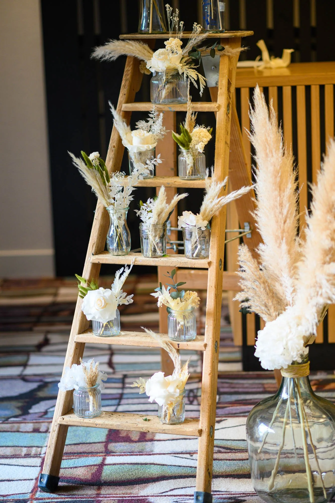 Une structure en bois avec plusieurs vases en verre contenant des fleurs blanches et pampas, décorations florales pour un événement