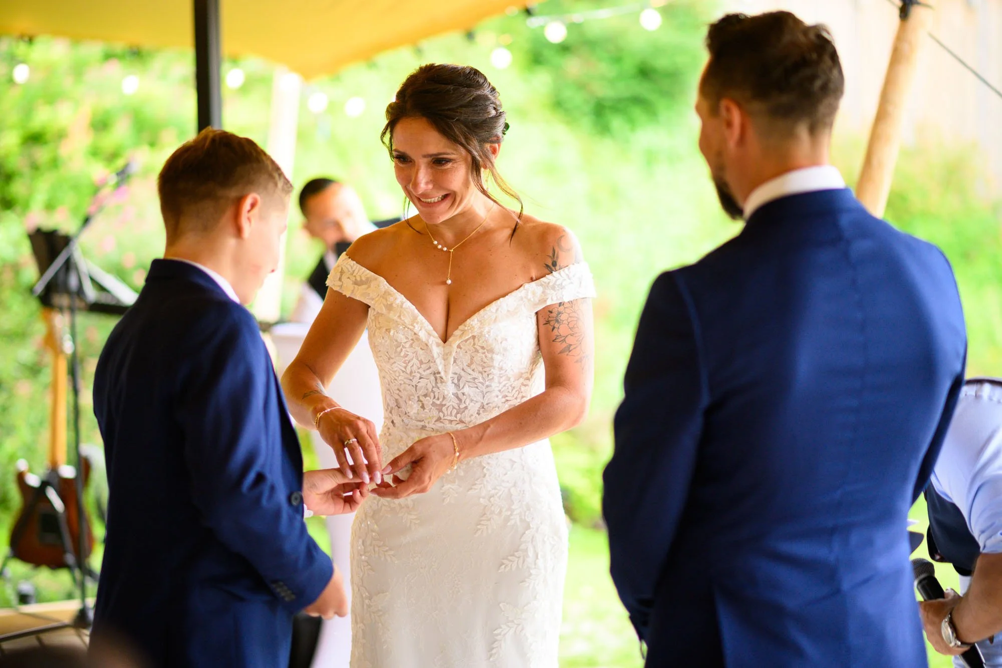 Une femme en robe de mariage échange des anneaux avec un garçon lors d'une cérémonie de mariage en plein air, entourée d'autres invités.