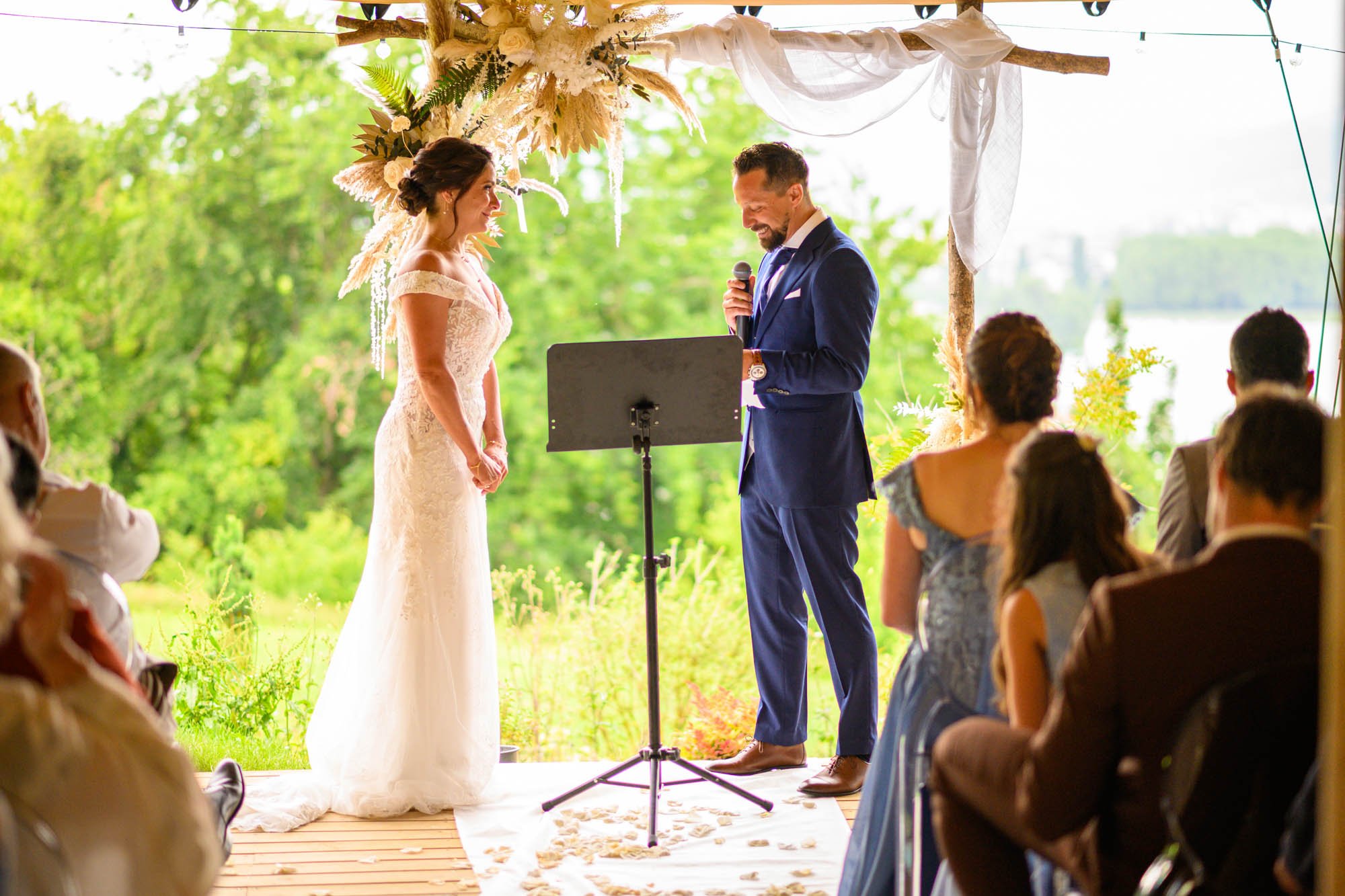 Un mariage en plein air avec la mariée en robe blanche et le marié en costume bleu. La cérémonie se déroule sous une Arche décorée de fleurs blanches et de feuillage, avec des invités assis regardant. Le pasteur ou l'officiant tient un micro et lit u