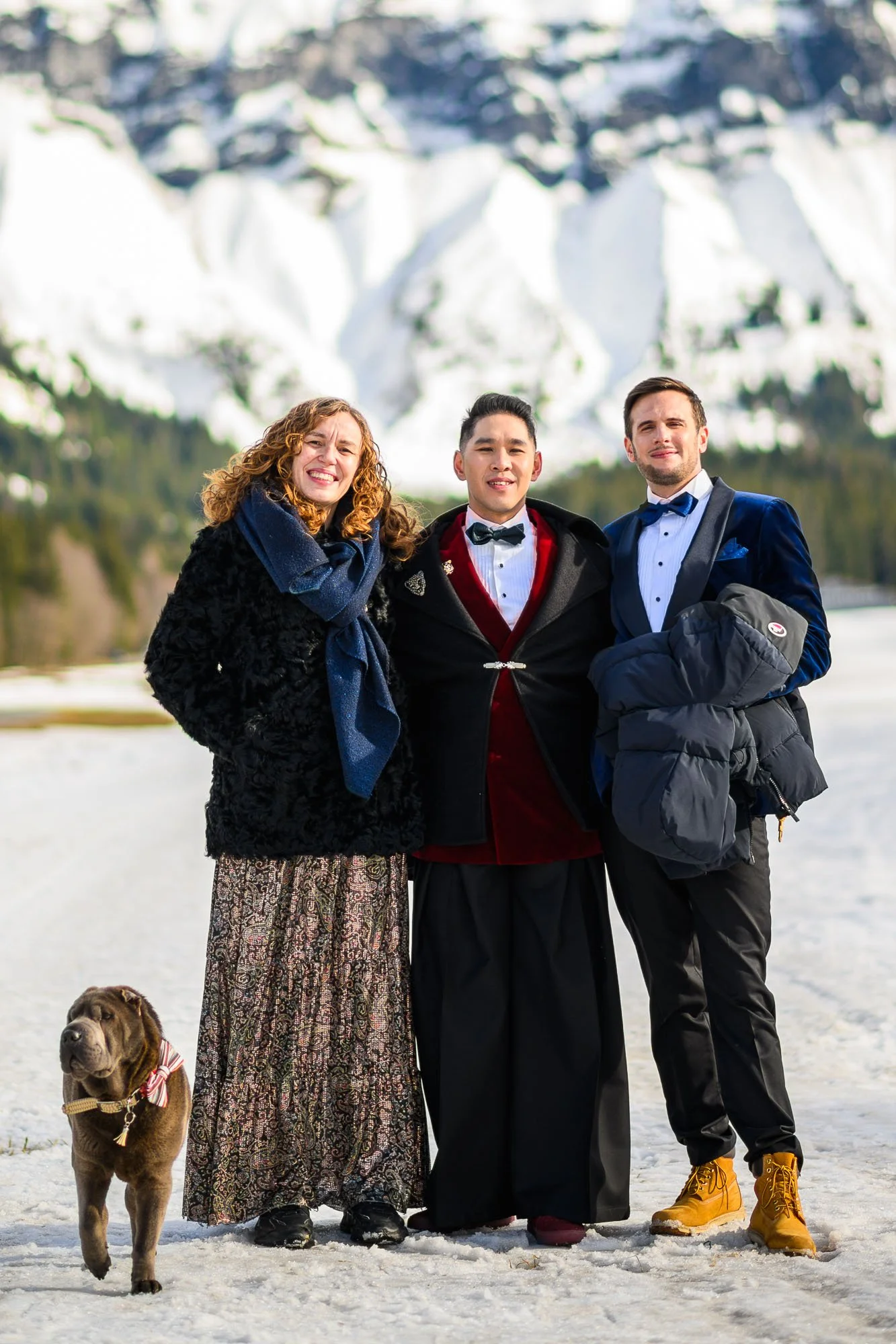 Trois personnes habillées formellement posent dans un paysage enneigé avec une montagne en arrière-plan, accompagnées d'un chien de petite taille.