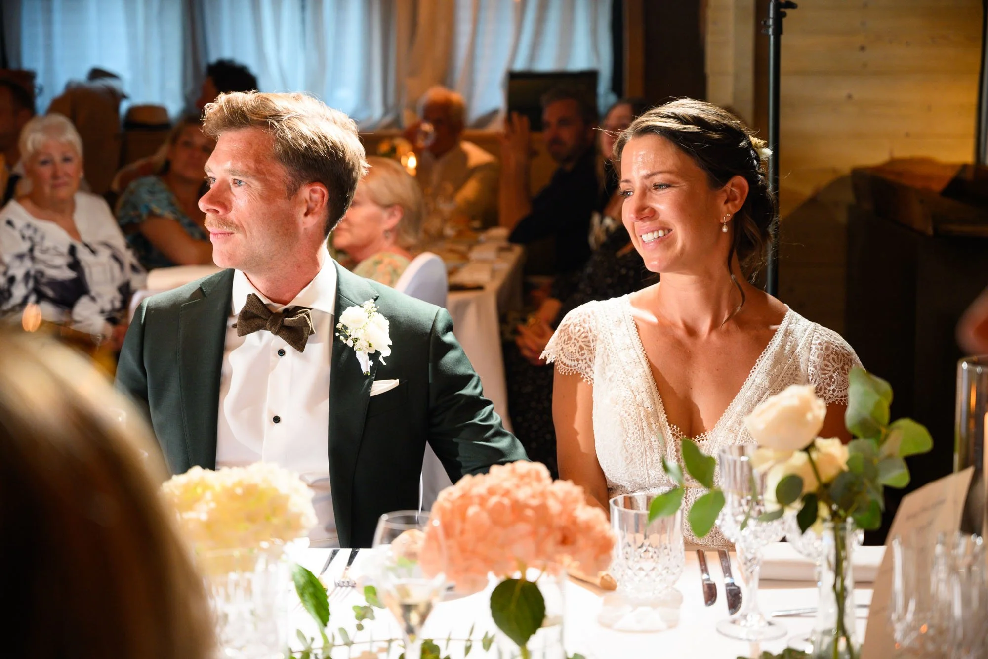 Un couple en tenue de mariage assis à une table lors d'une réception, entouré d'invités.