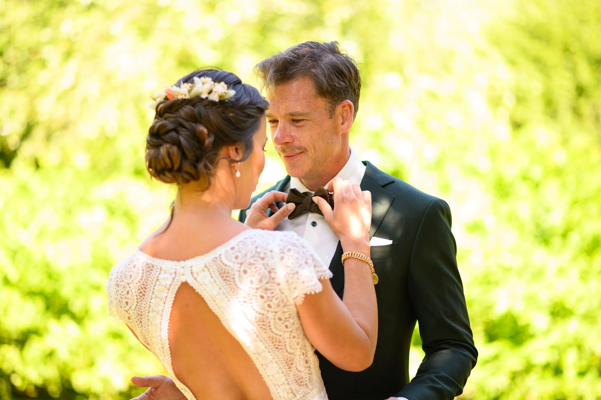 Un couple de mariés en pleine séance photo en plein air. La mariée ajuste le nœud papillon du marié, tous deux souriants, dans un environnement verdoyant, ensoleillé.