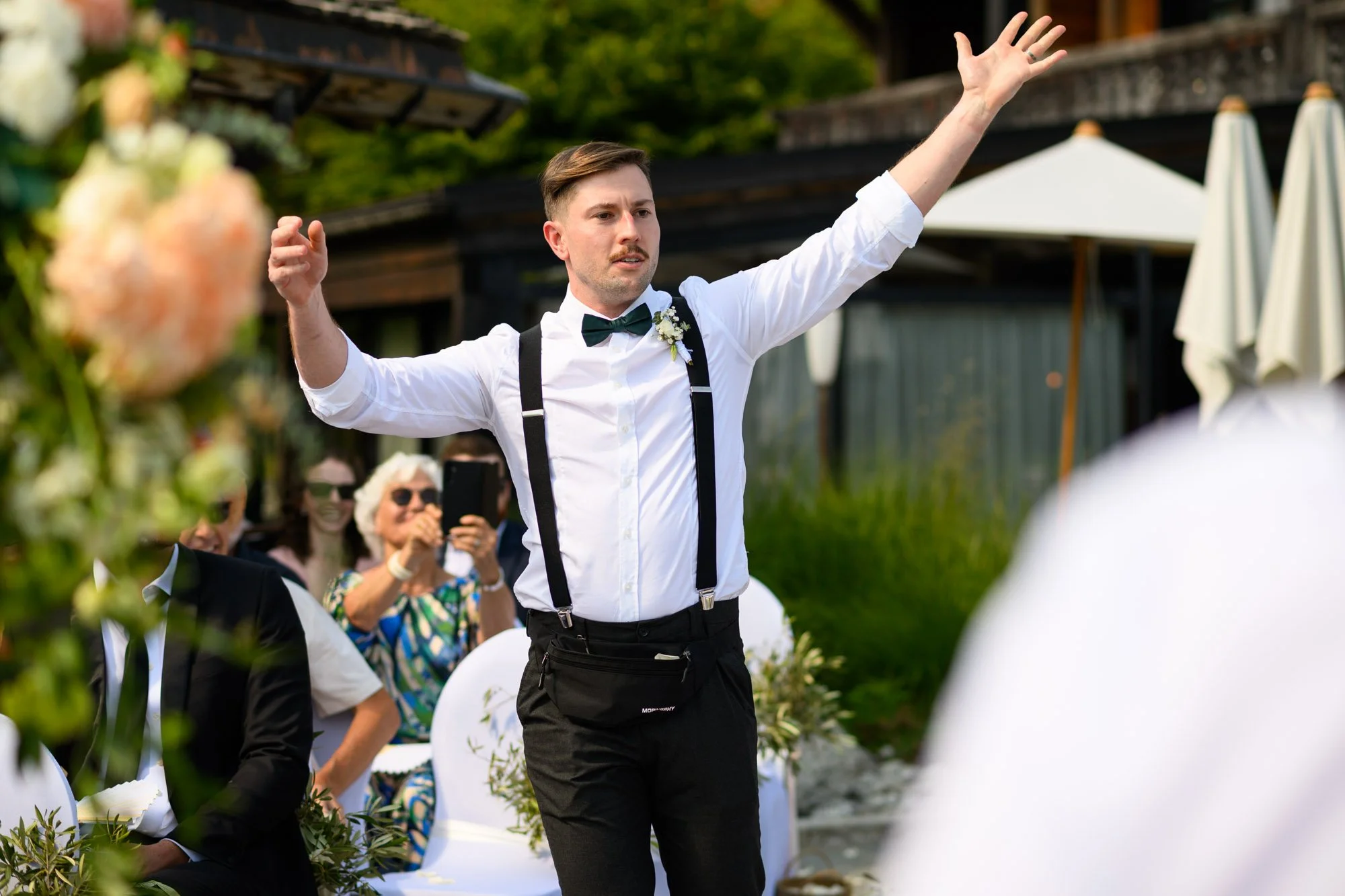 Un homme danse lors d'un mariage en extérieur, portant une chemise blanche, un nœud papillon vert, et des bretelles noires, avec des invités qui regardent et prennent des photos.