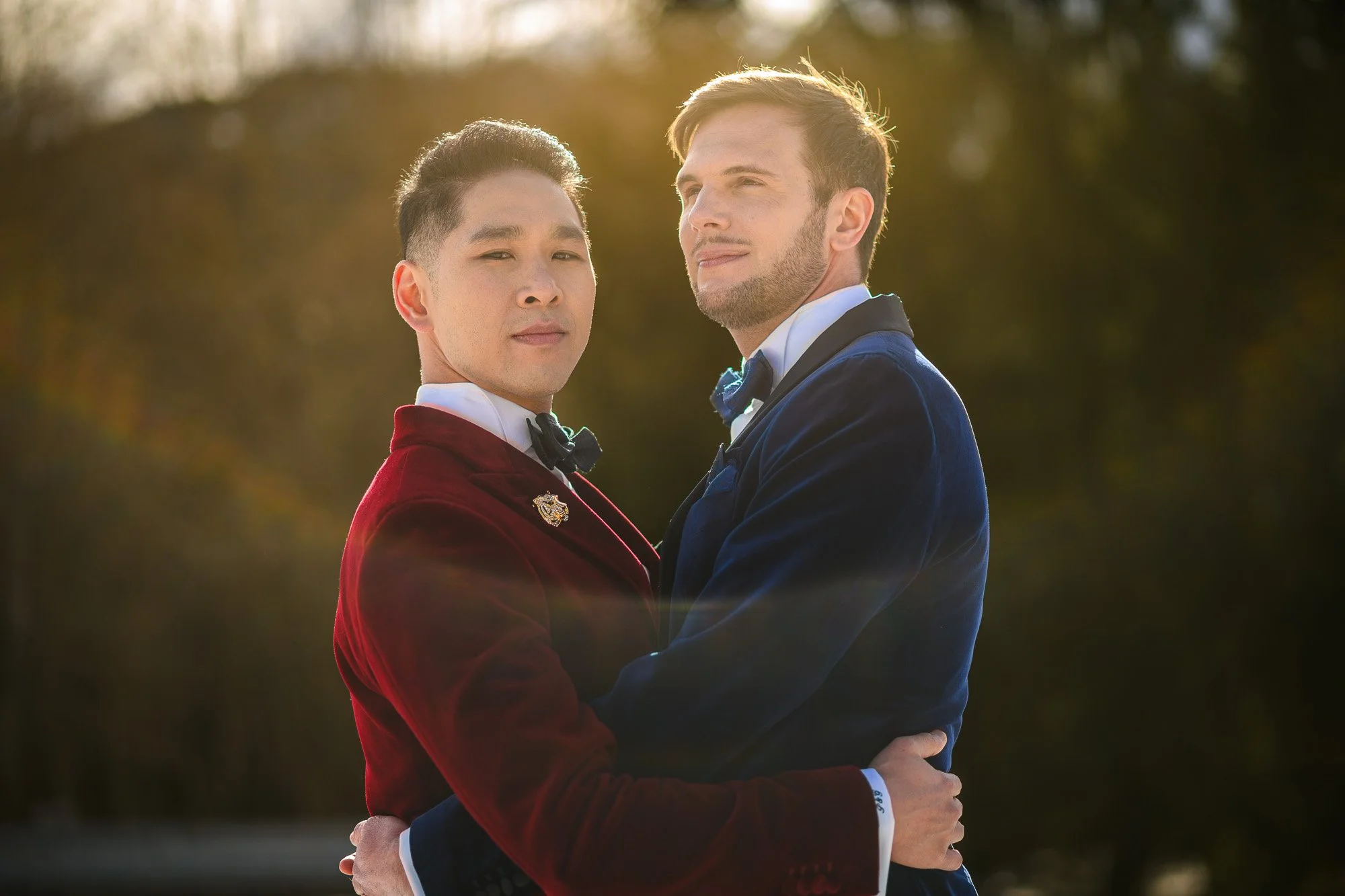 Deux hommes en costume de soirée se tiennent dans un parc avec des arrière-plans flous de foliage; ils se tiennent l'un face à l'autre, se tenant la taille.