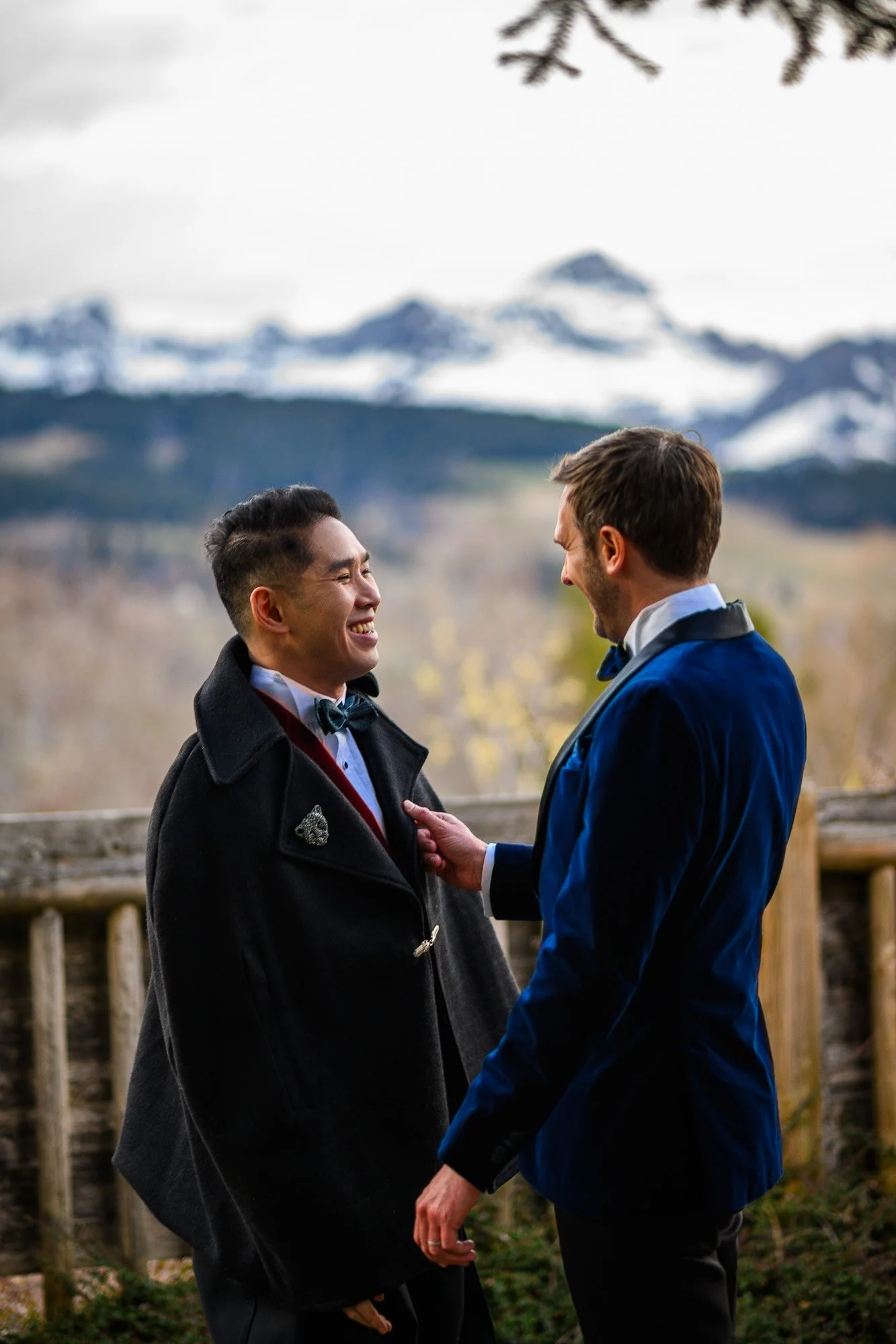 Deux hommes habillés en costumes de mariage, partageant un moment de bonheur avec des montagnes enneigées en arrière-plan.